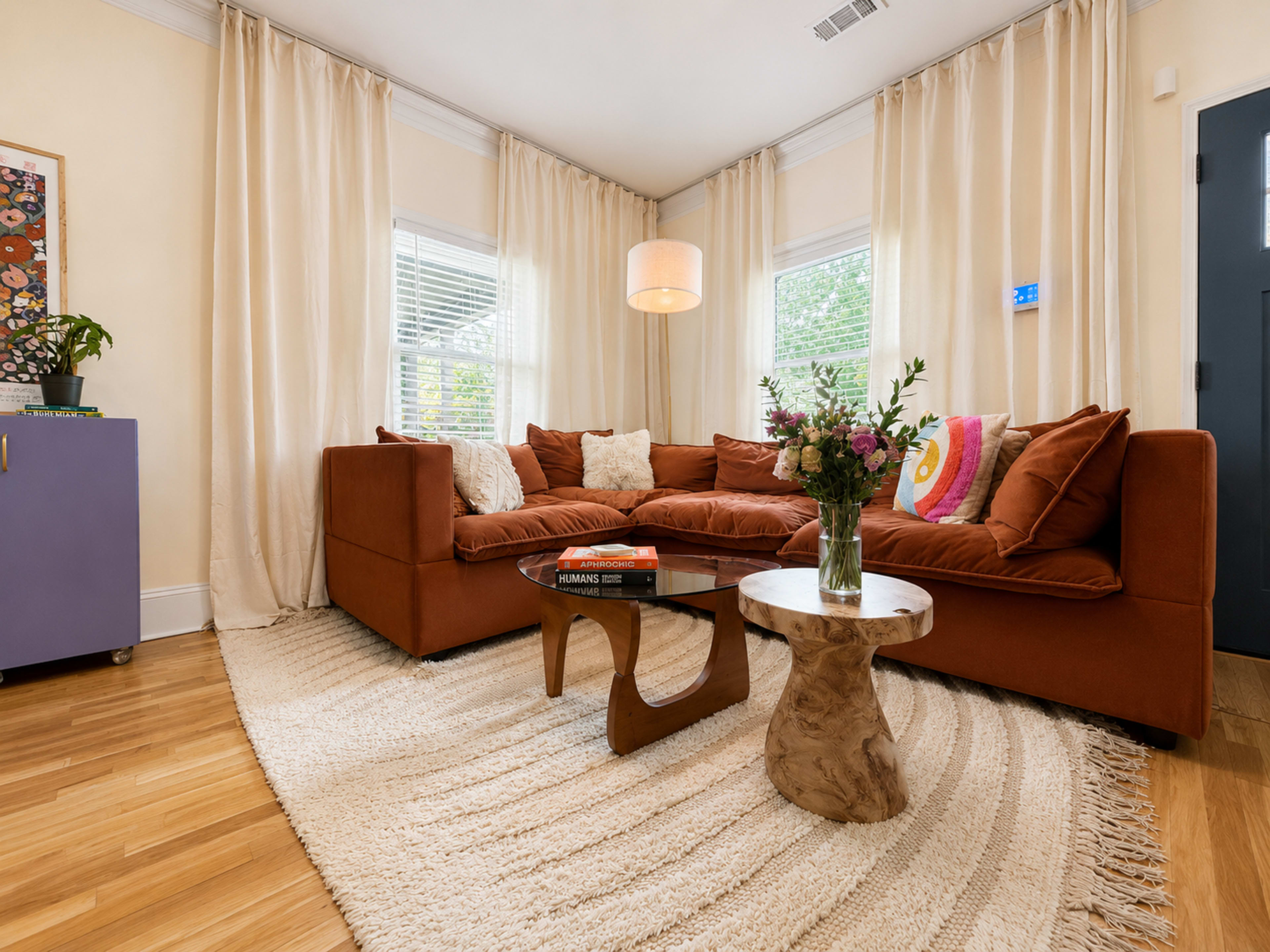The image shows a cozy living room featuring a large sectional sofa, a small wooden side table, a vase of flowers, and light-colored curtains.