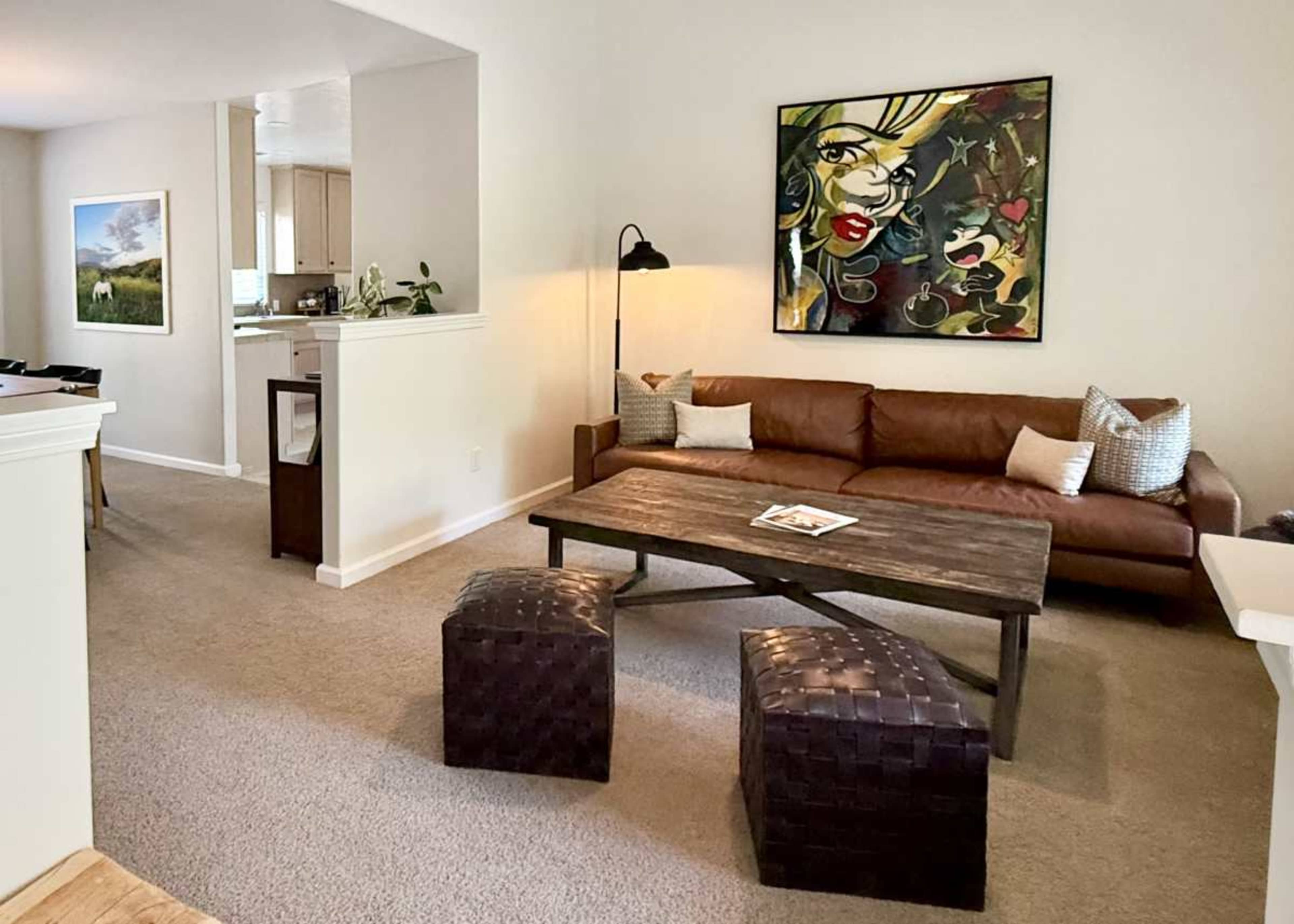 The image shows a living room featuring a brown leather sofa, a wooden coffee table, and decorative woven stools, with a colorful painting hanging on the wall.