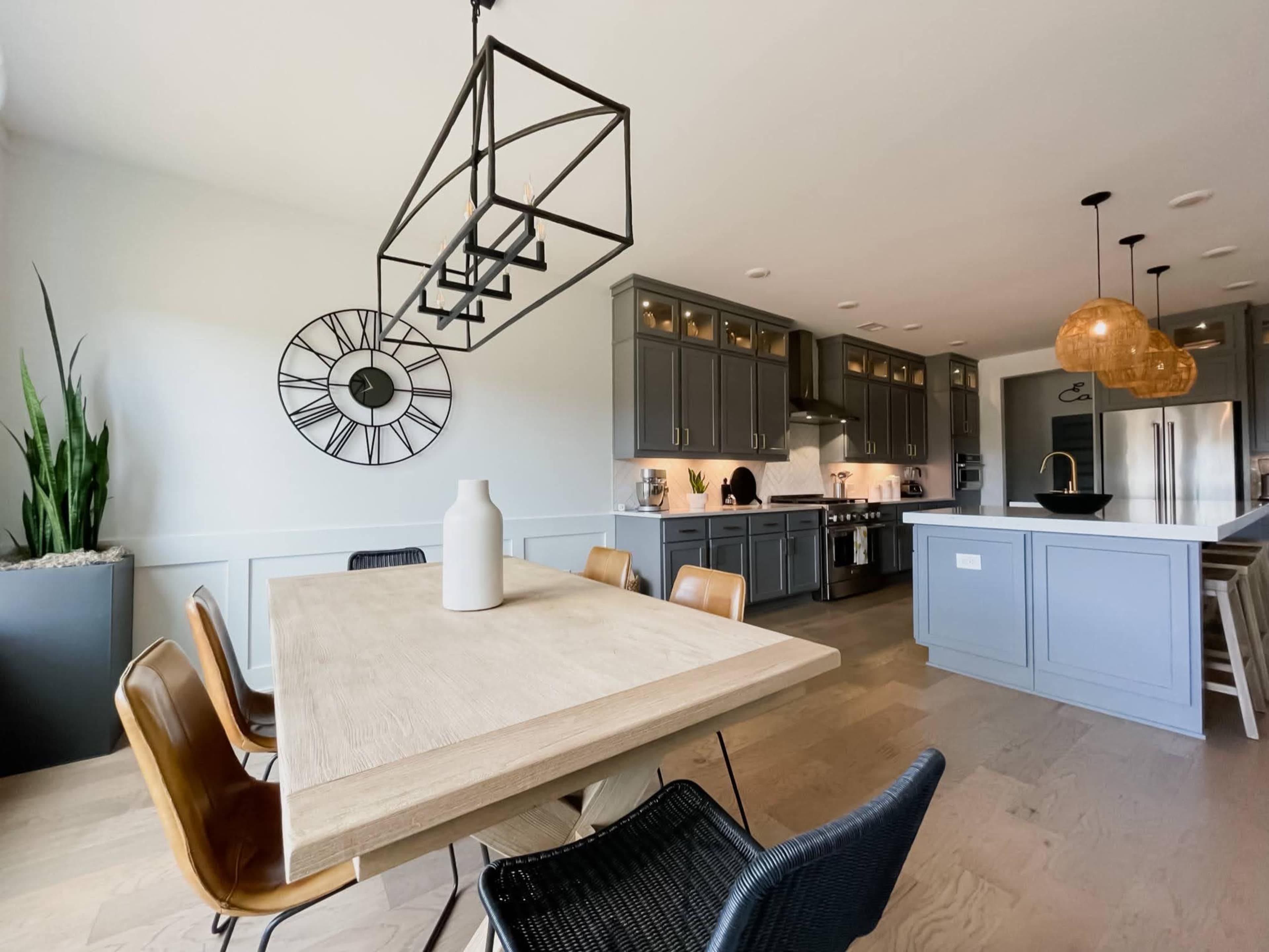 A modern kitchen features gray cabinetry, a large wooden dining table, and pendant lighting above the island.