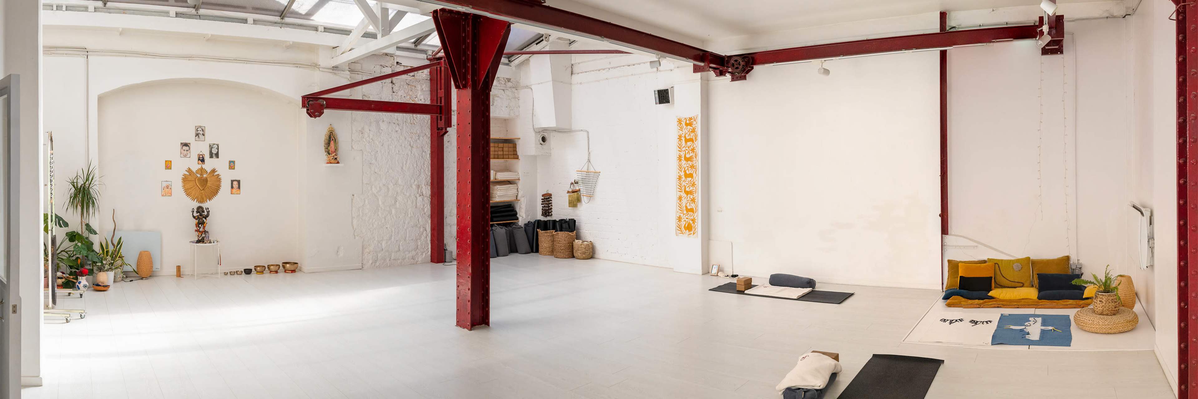 The image shows a spacious yoga studio with white walls, wooden flooring, and various mats and cushions arranged for practice.