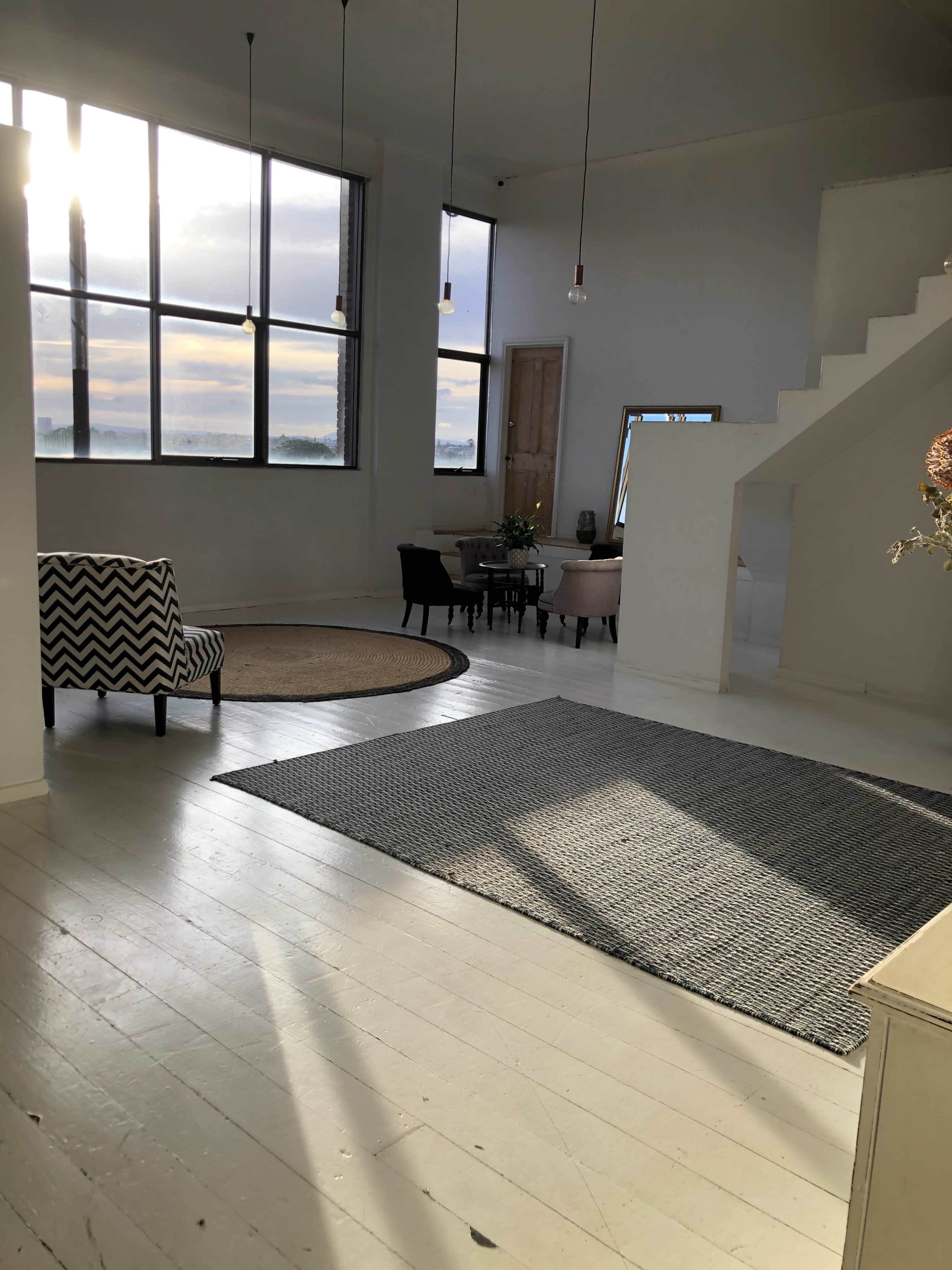 A spacious, modern interior with large windows, a patterned chair, a round area rug, and a staircase leading to a second level.