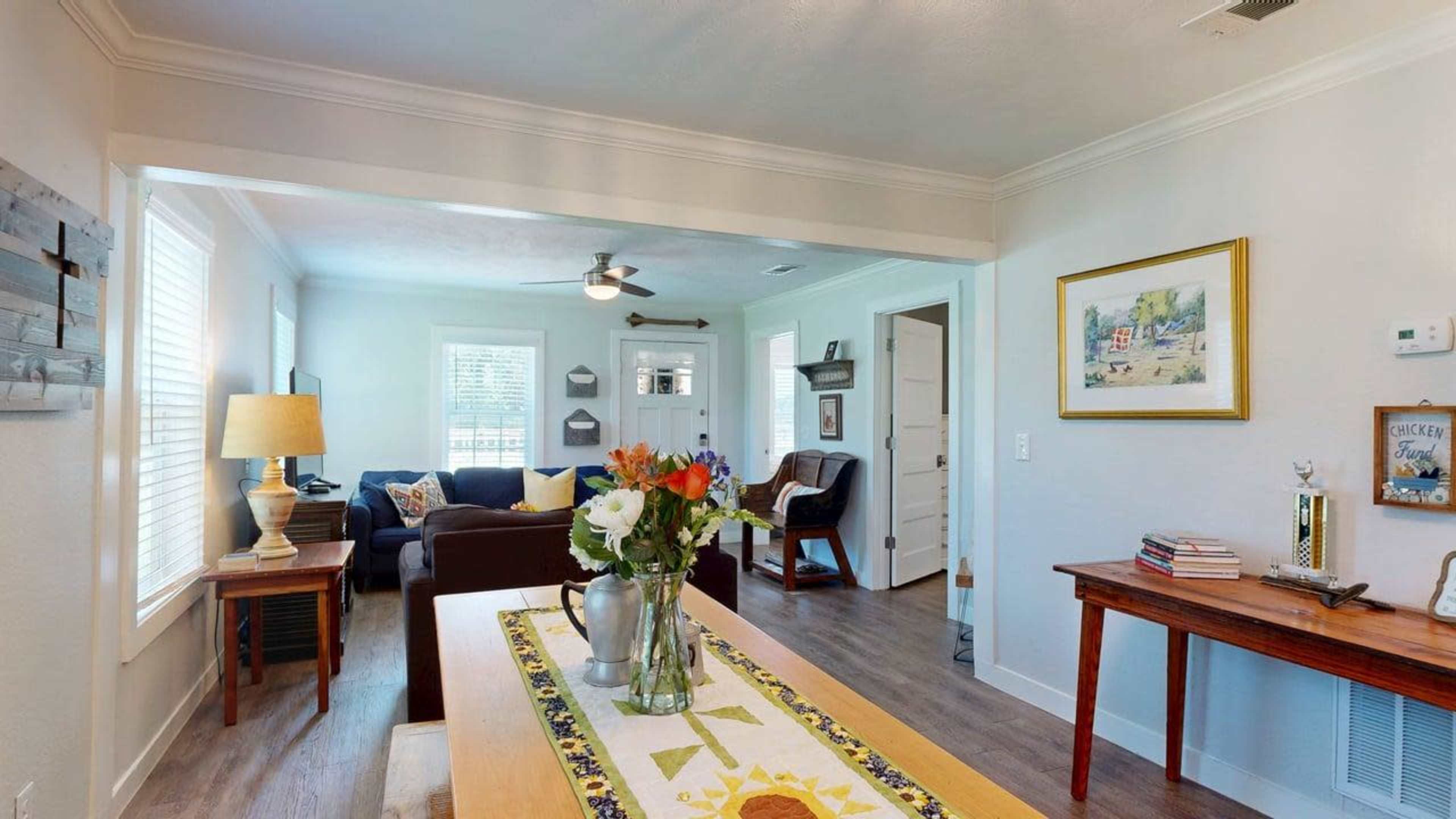 The image shows a cozy living space that features a dining table with a floral centerpiece, a seating area with a sofa and chair, and a well-lit atmosphere from windows and light fixtures.