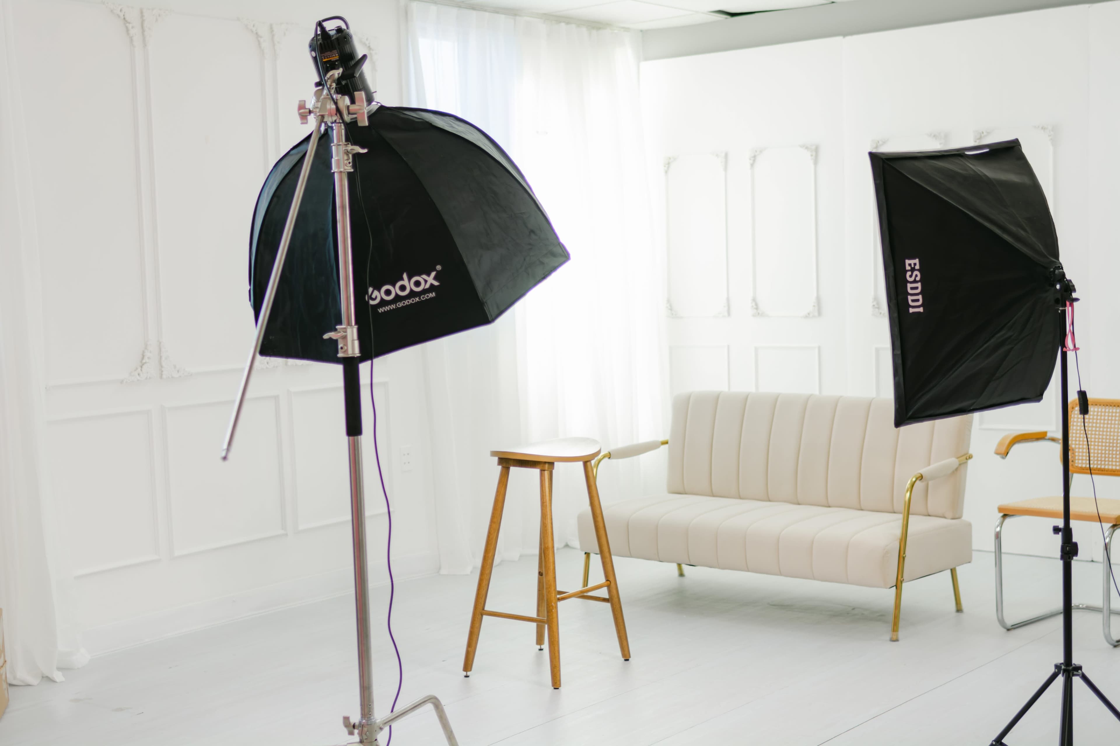 The image shows a photography studio with two softbox light setups, a wooden stool, and a beige couch against a white wall.