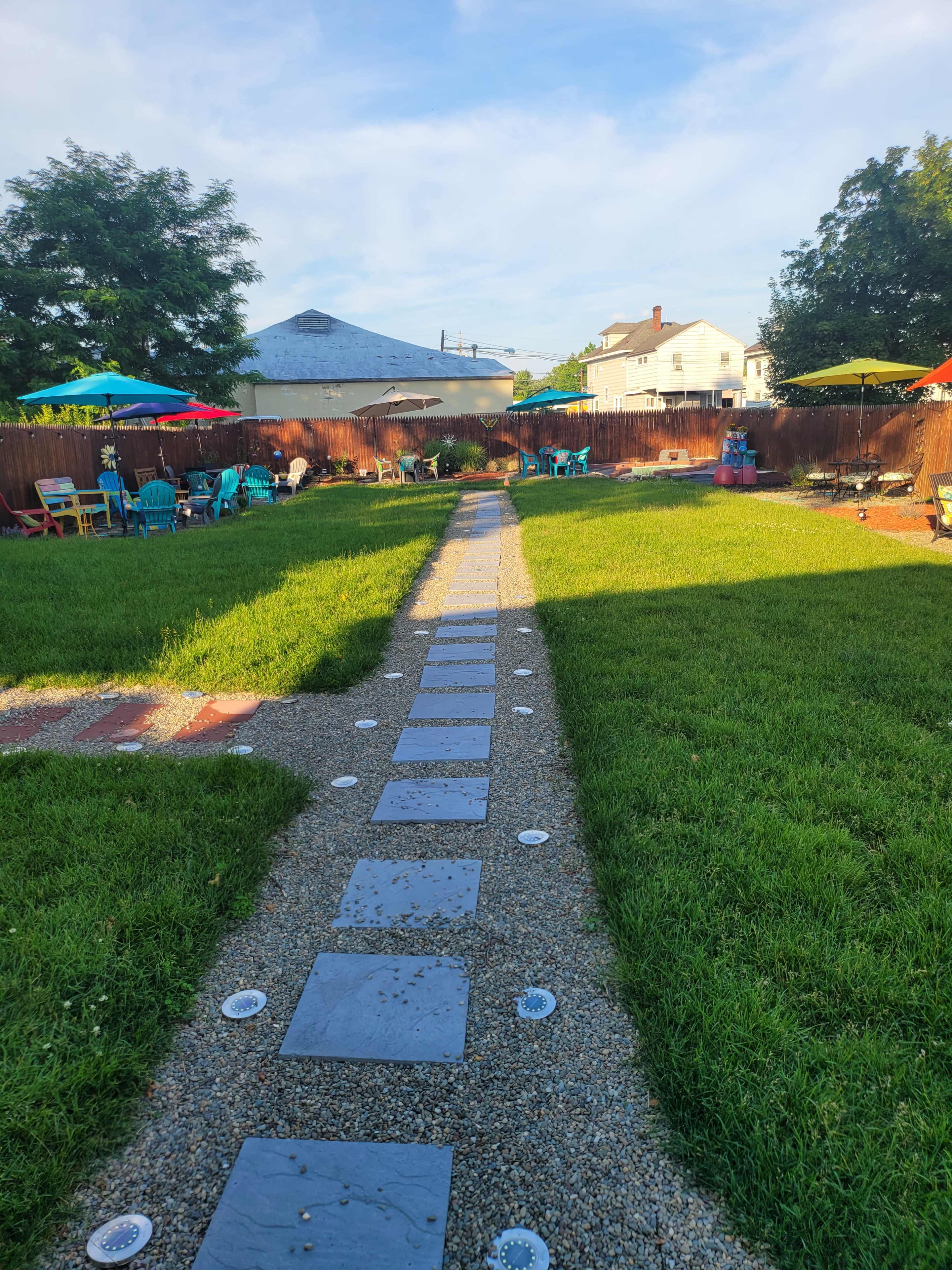 A stone pathway leads through a grassy backyard filled with colorful seating areas and shaded umbrellas.