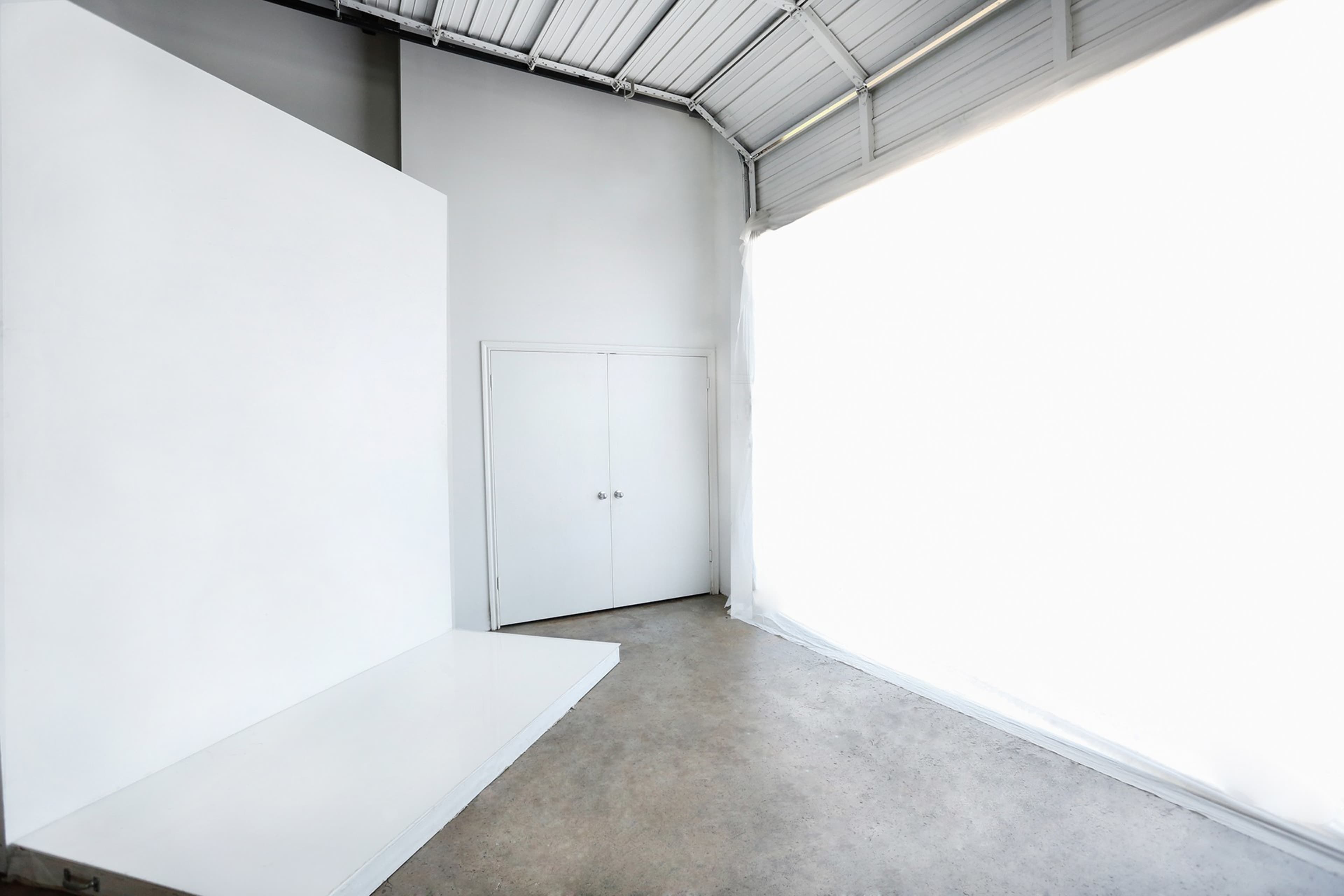 A spacious, minimalist room features a large white backdrop and a double door on one wall, with natural light flowing in from an expansive window.