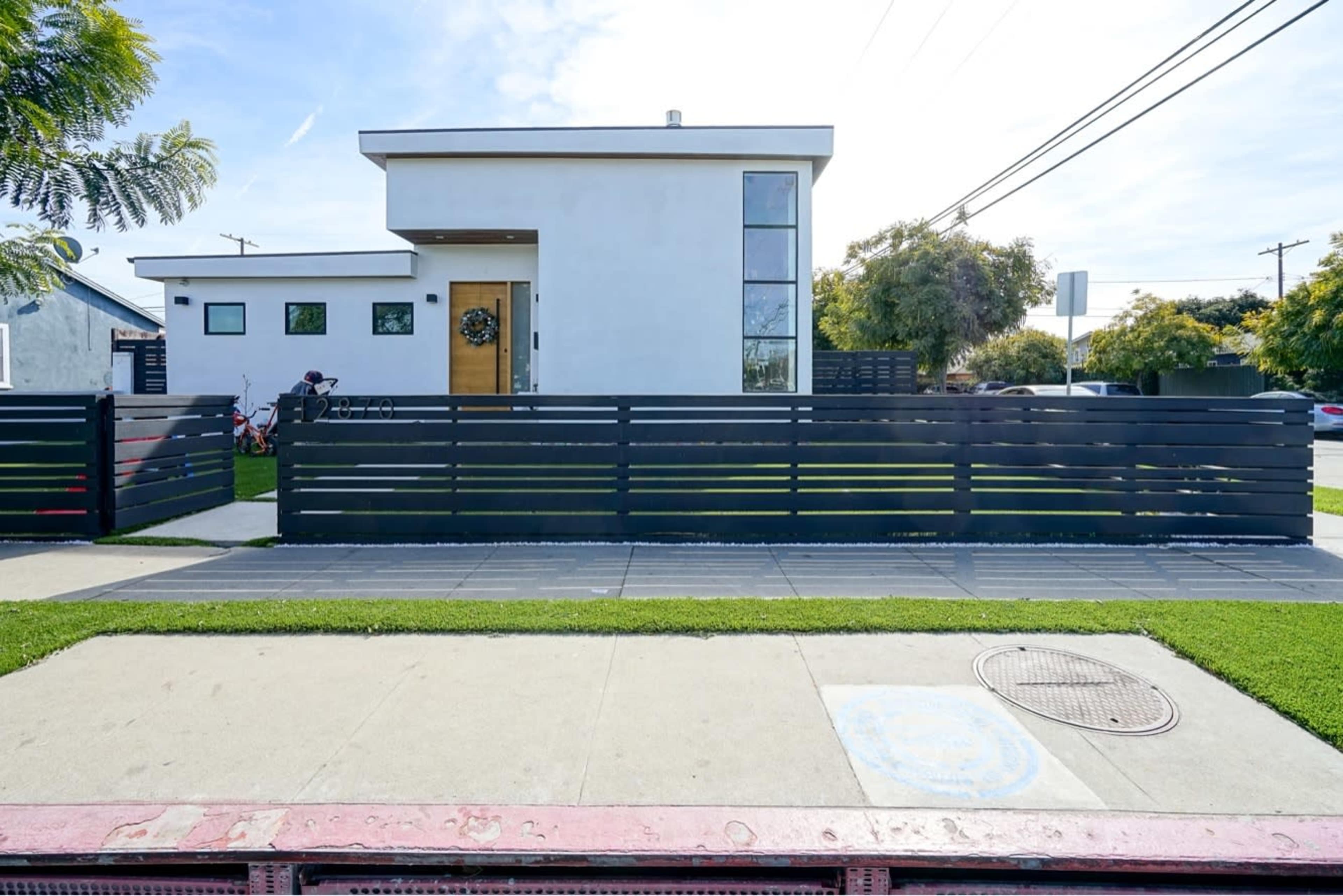 A modern white house with large windows and a wooden fence is situated on a landscaped front yard alongside a sidewalk.