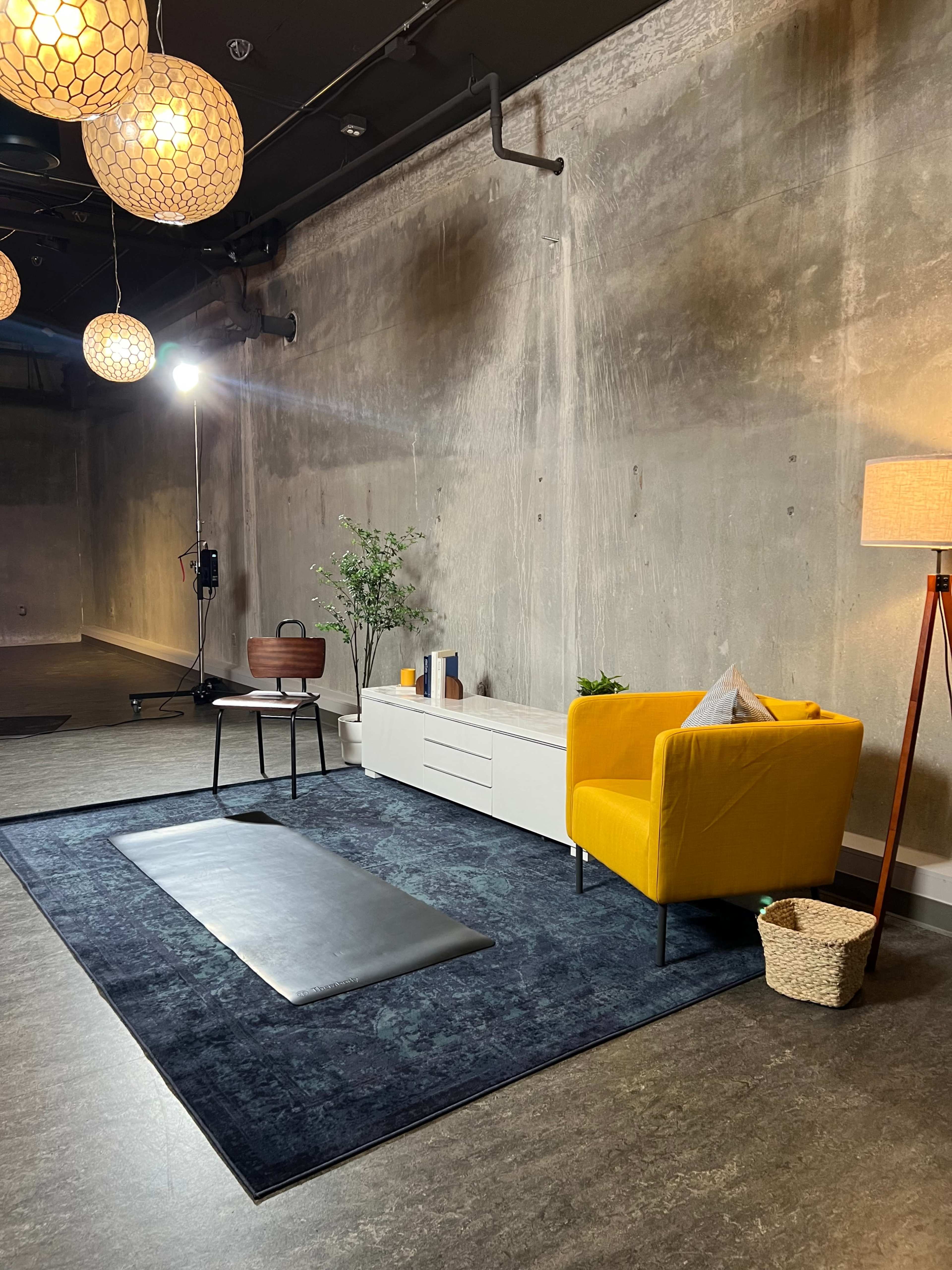 A modern interior space with a yellow armchair, a black yoga mat on the floor, a white storage unit, and a lamp against a concrete wall.