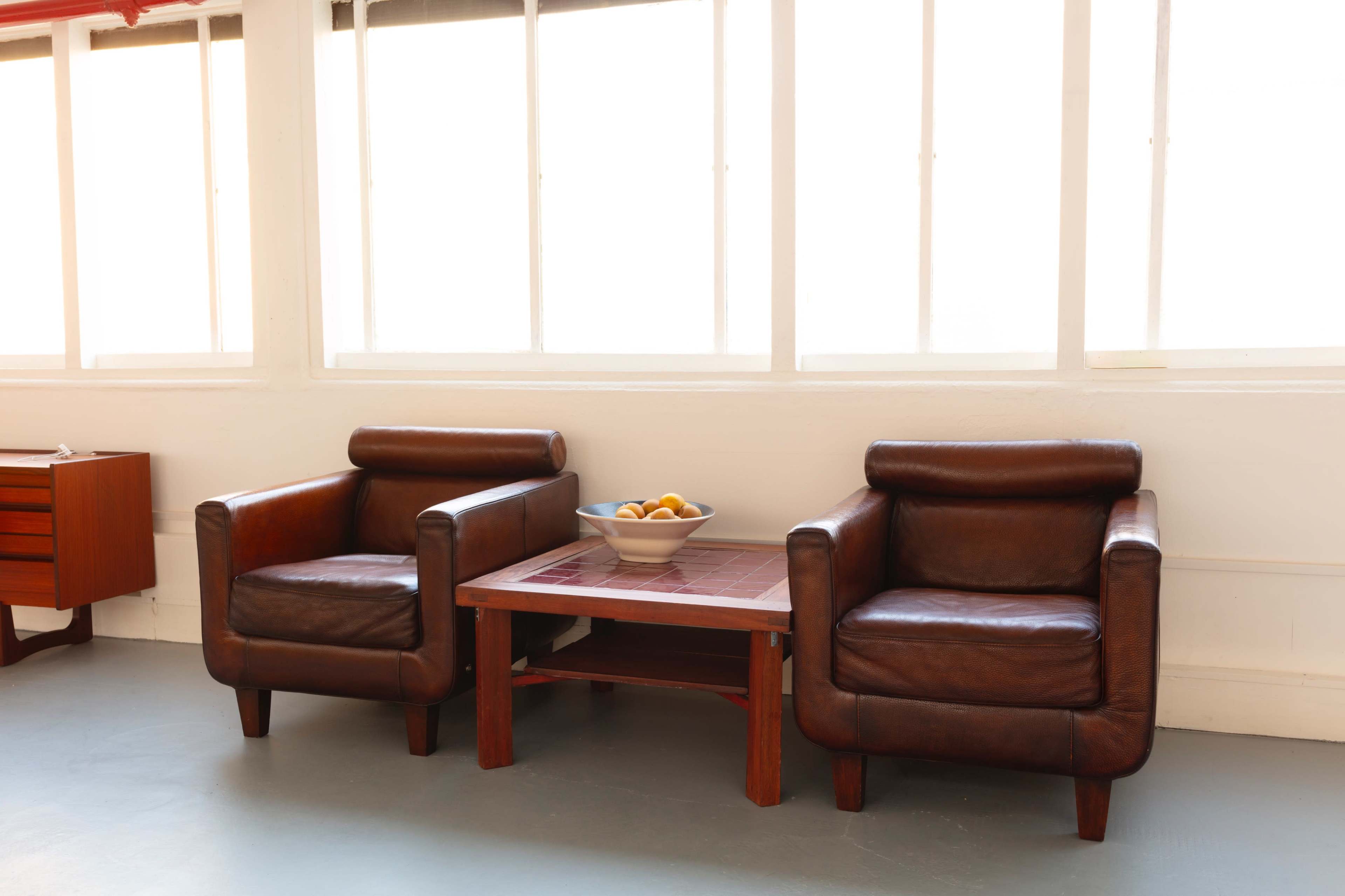 Two brown leather armchairs are placed opposite each other around a wooden coffee table with a bowl of fruit on it, in a brightly lit room with large windows.