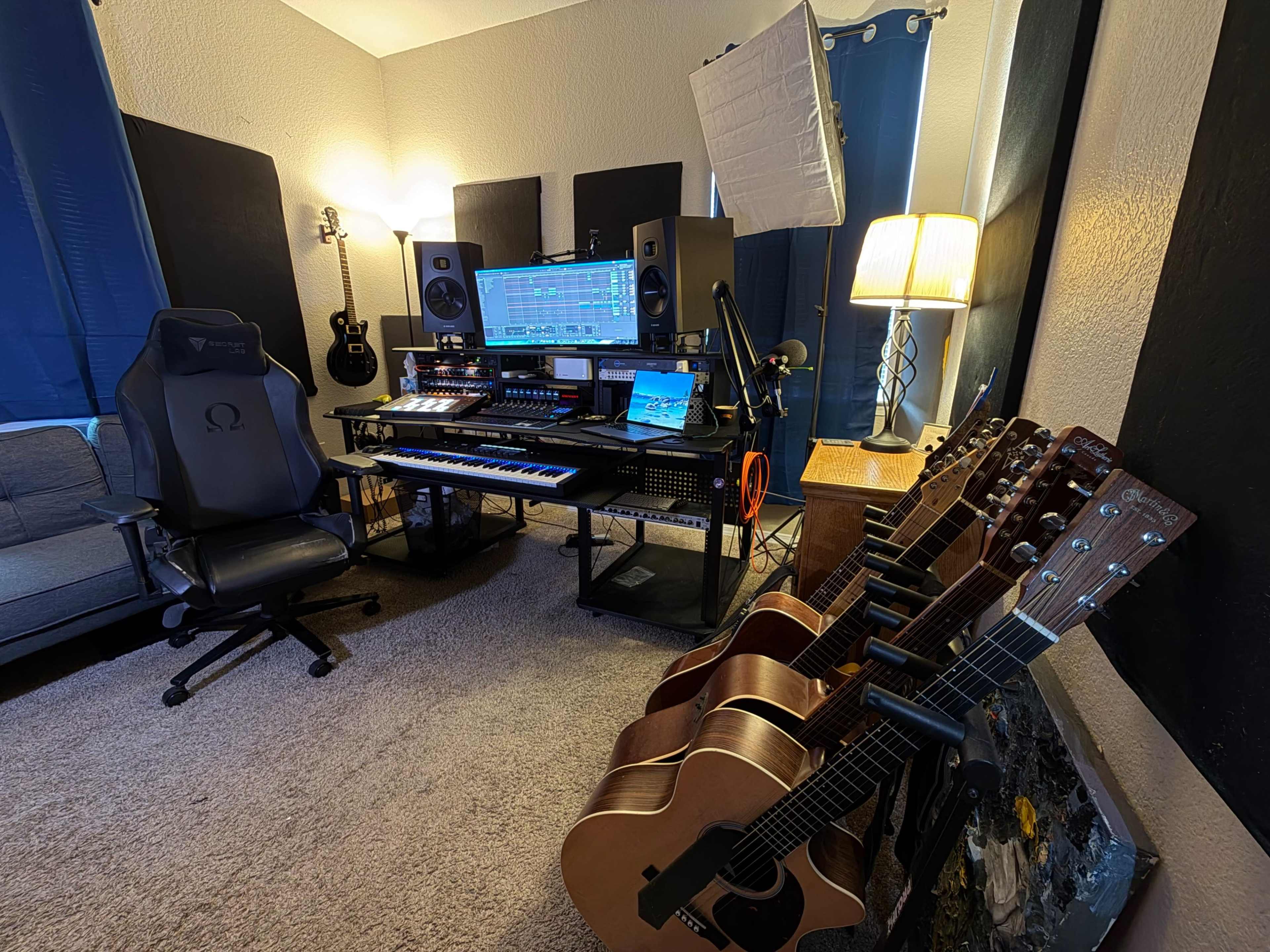 The image shows a music recording studio featuring a keyboard, computer monitors, audio equipment, a microphone, and several guitars arranged neatly in a corner.