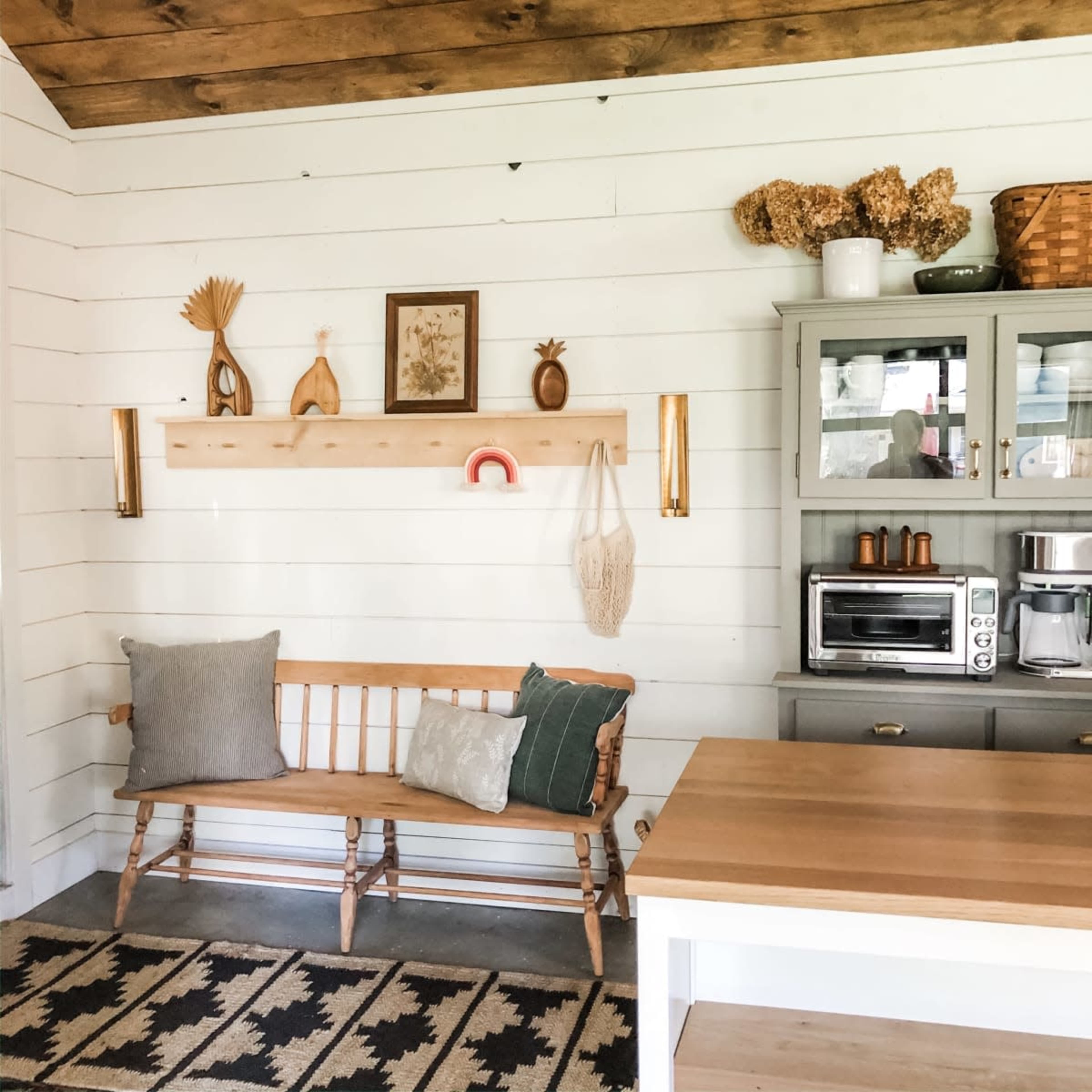 A cozy kitchen area features a wooden bench against a white wall, adorned with decorative items and a gray cabinet displaying kitchen appliances.