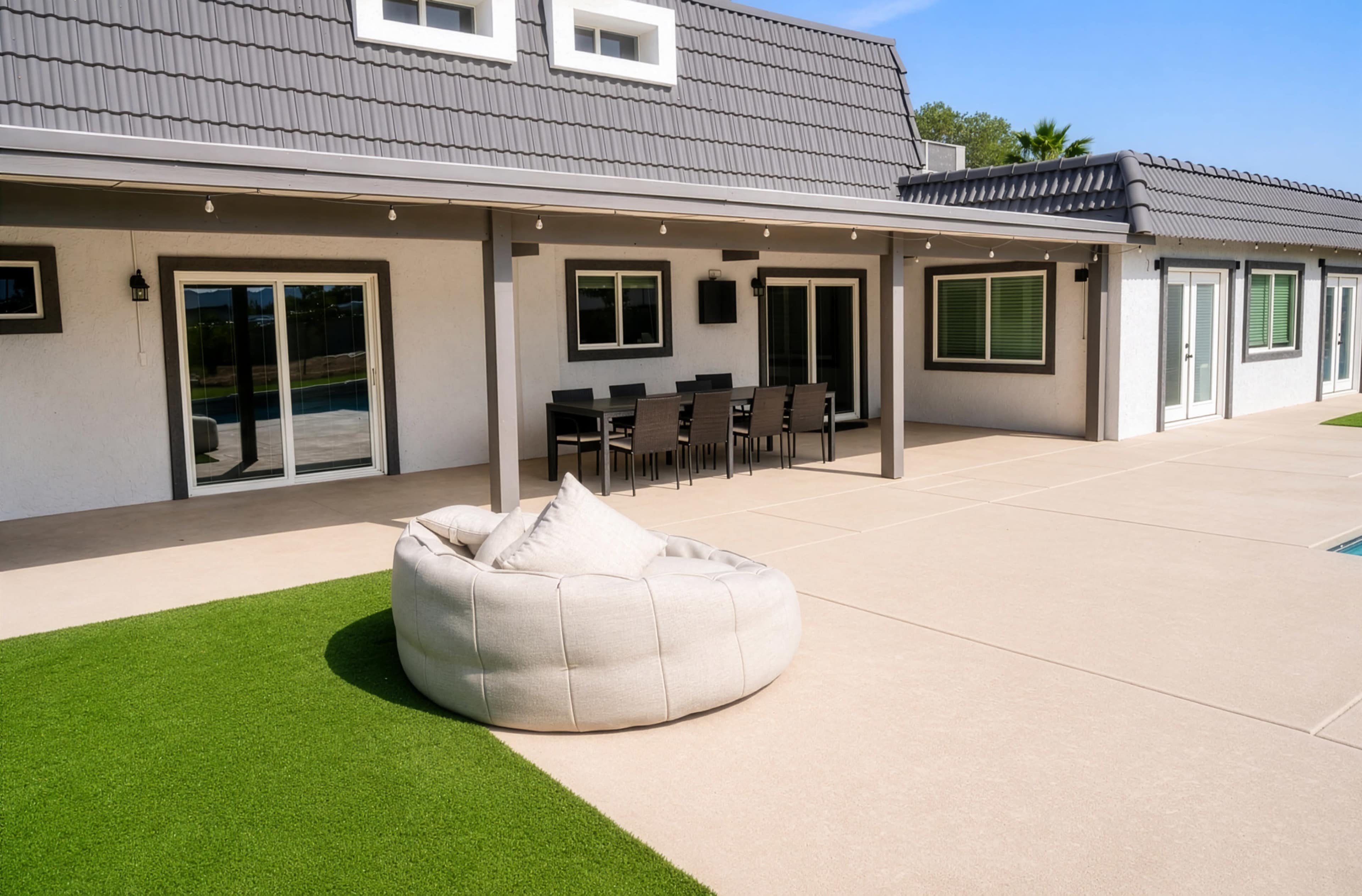 The image shows a spacious outdoor patio area featuring a seating arrangement with a dining table, a large cushioned beanbag, and artificial grass.