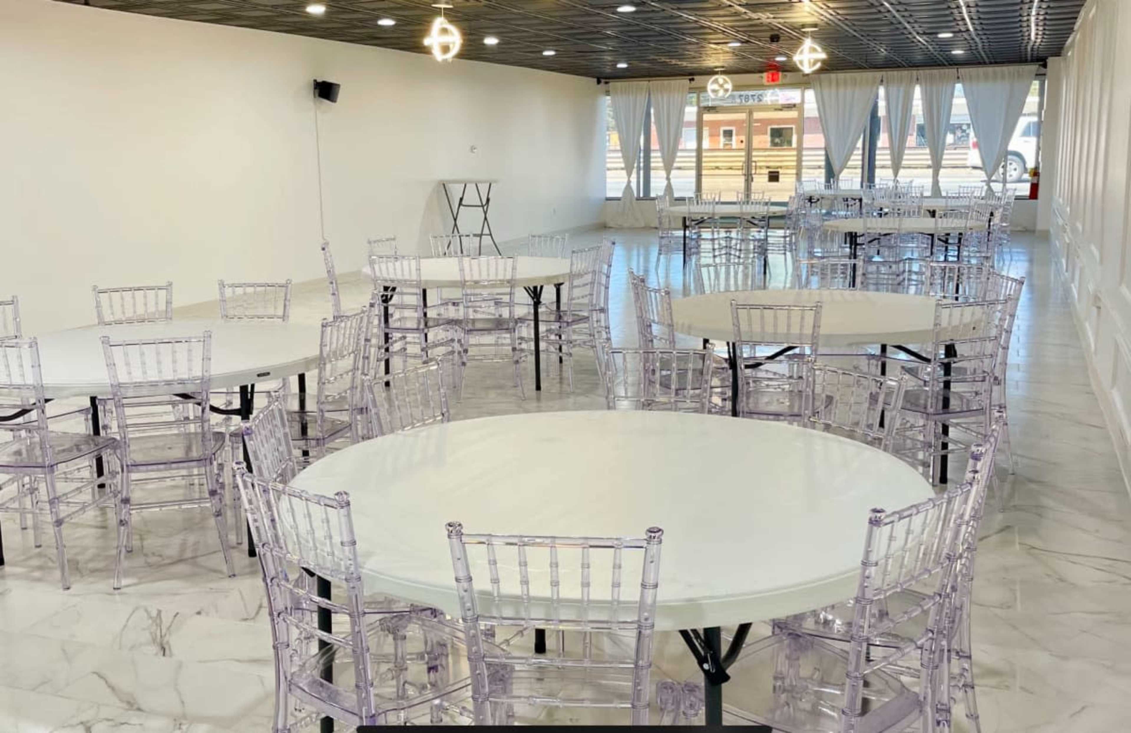 A spacious event hall with multiple round tables surrounded by transparent chairs and large windows allowing natural light.