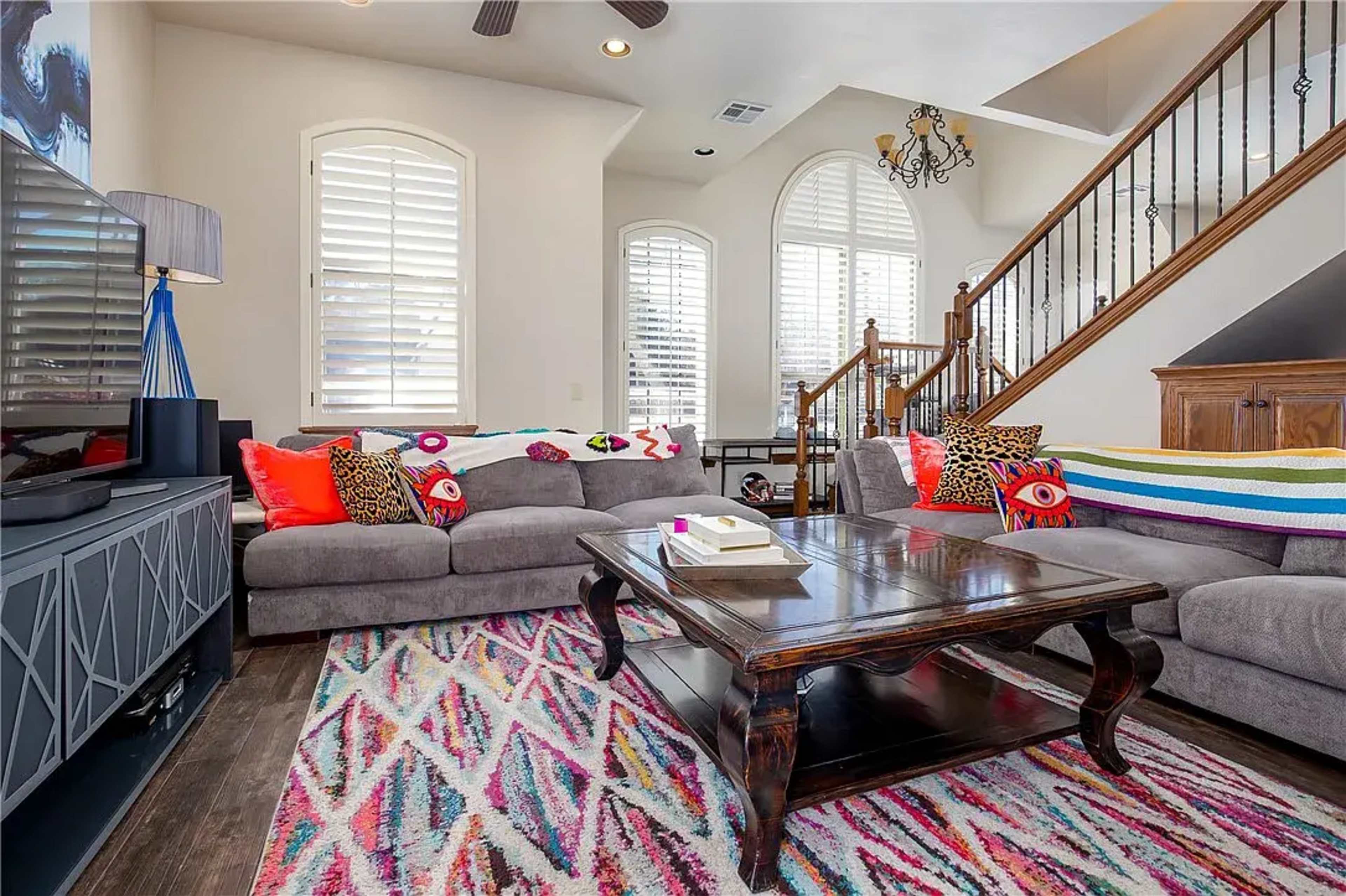 The living room features a spacious layout with two gray sofas adorned with colorful pillows, a dark wooden coffee table, and a patterned area rug, all complemented by large windows and a staircase in the background.