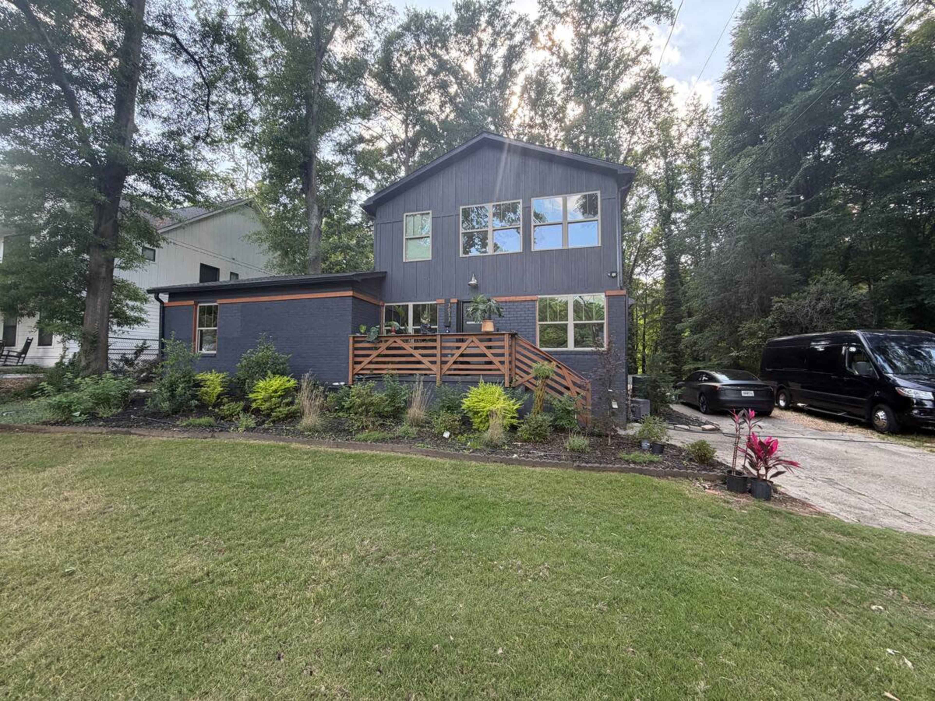 A modern two-story house with a dark exterior and large windows is surrounded by greenery and has a wooden staircase leading to the entrance.
