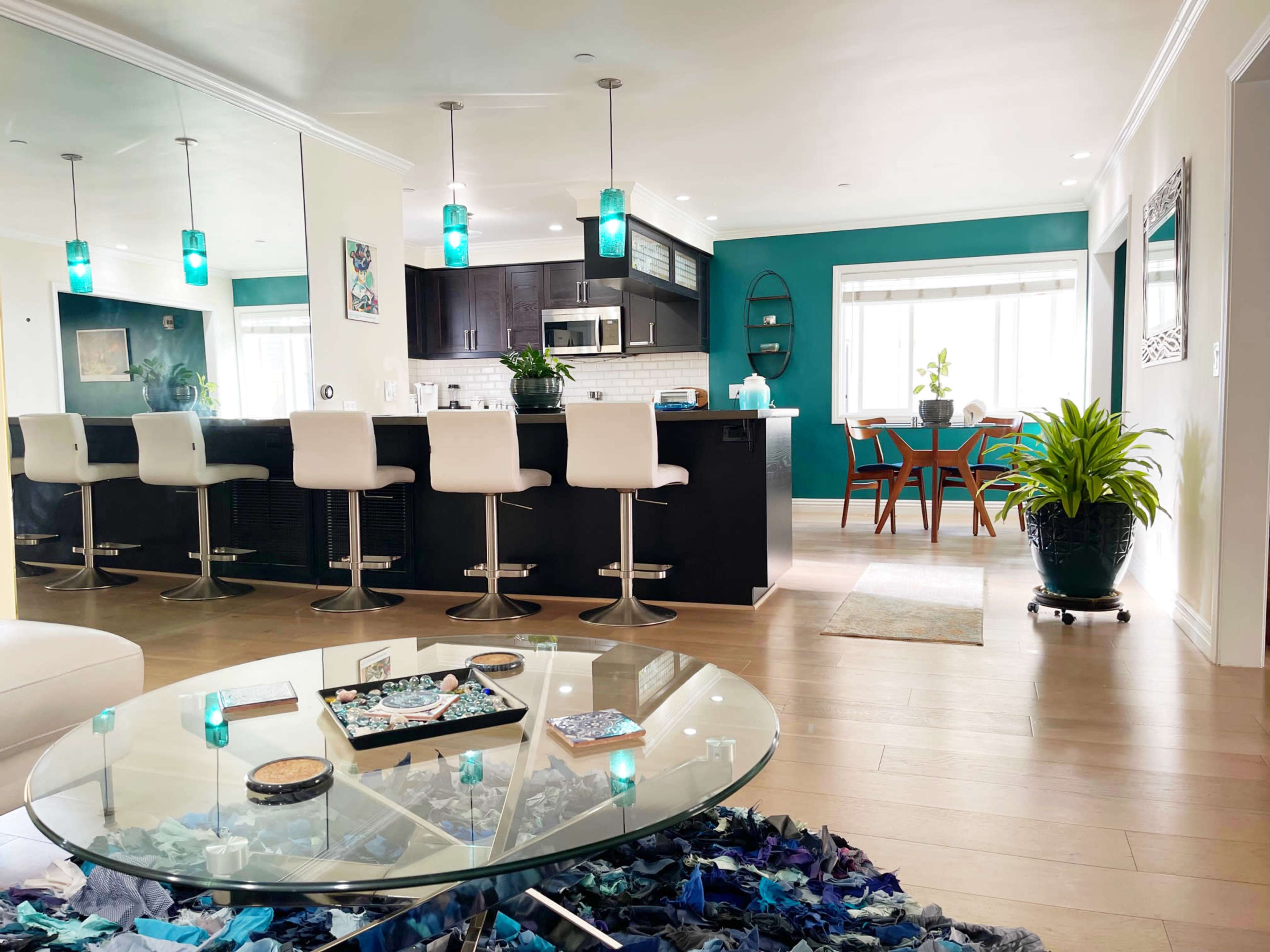 The image shows a modern open-concept living space featuring a kitchen with dark cabinetry, a bar with high stools, and a dining area with a large window.