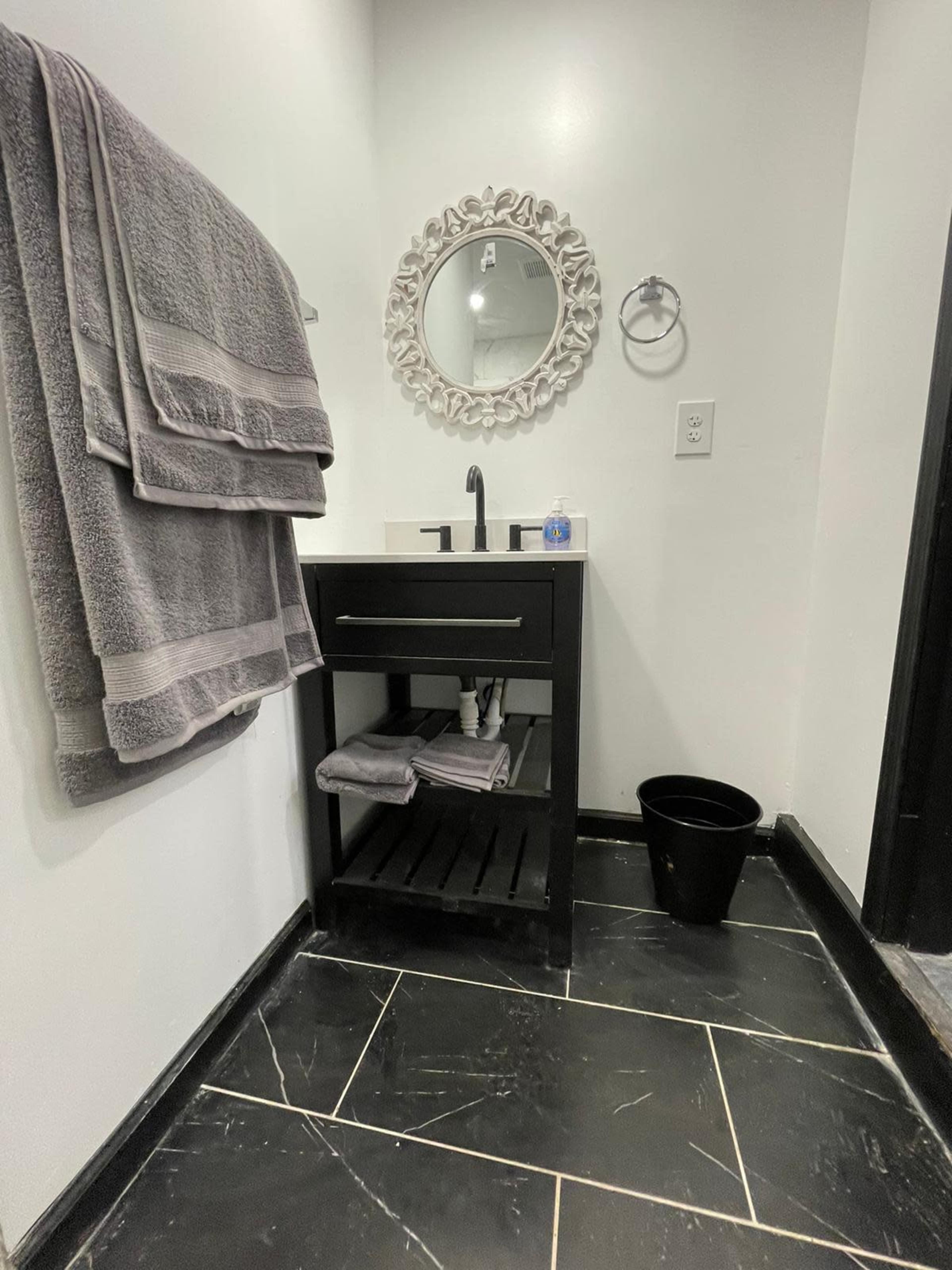 A small bathroom featuring a black vanity with a sink, a round decorative mirror, gray towels hanging on the wall, and a black wastebasket on the floor.