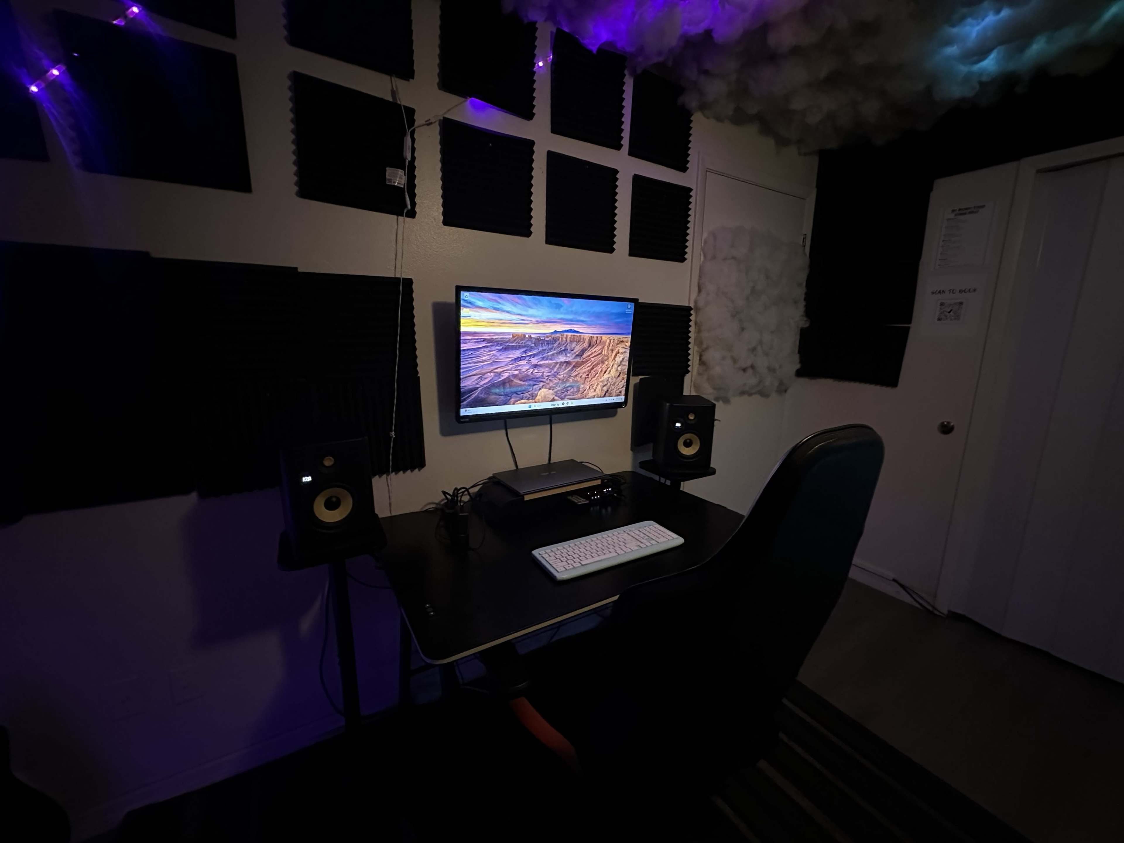 The image shows a desk setup with a monitor, speakers, and a keyboard against a wall adorned with soundproofing panels and artificial clouds.