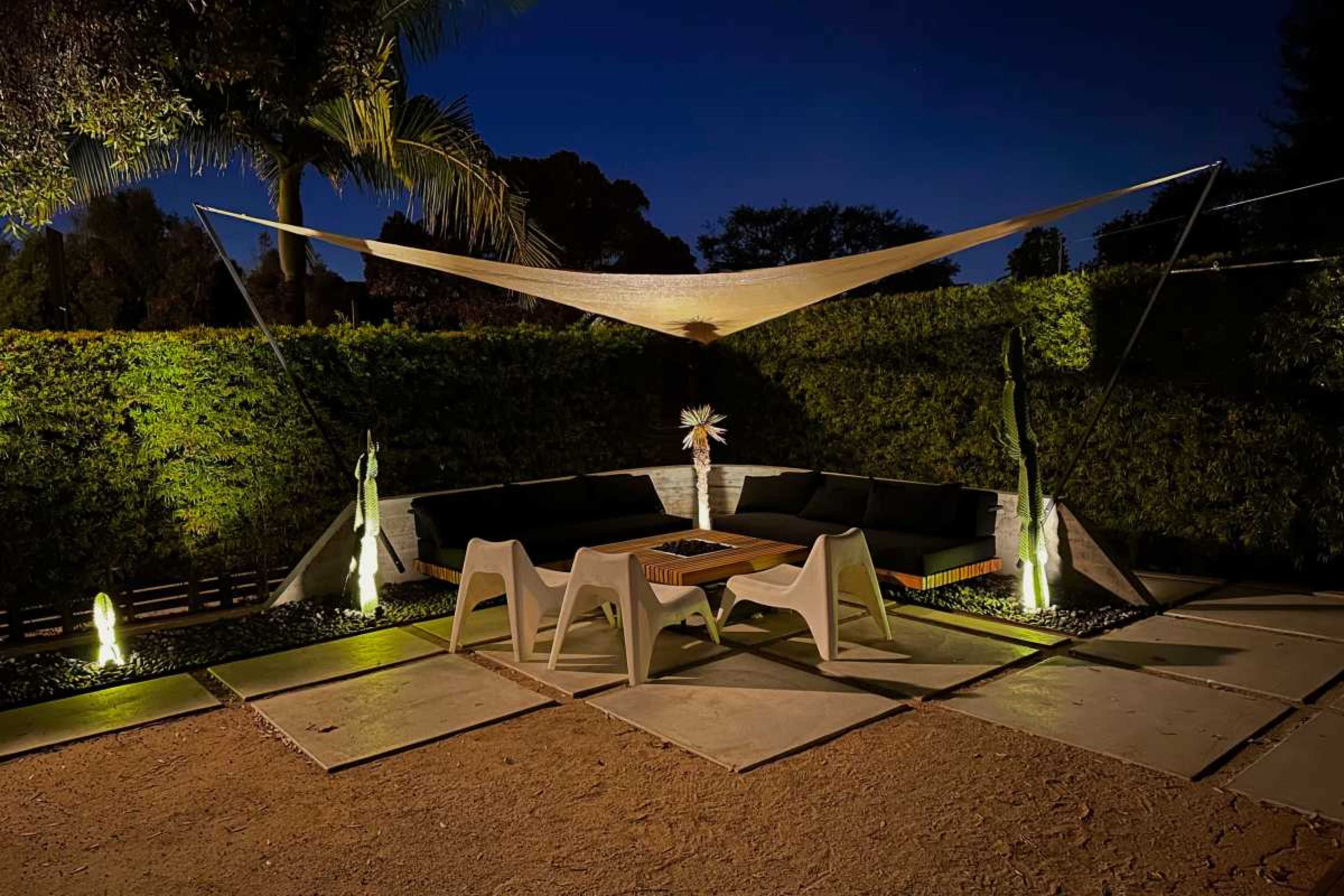 A modern outdoor seating area features a shaded lounge couch and a coffee table, surrounded by greenery under nighttime illumination.