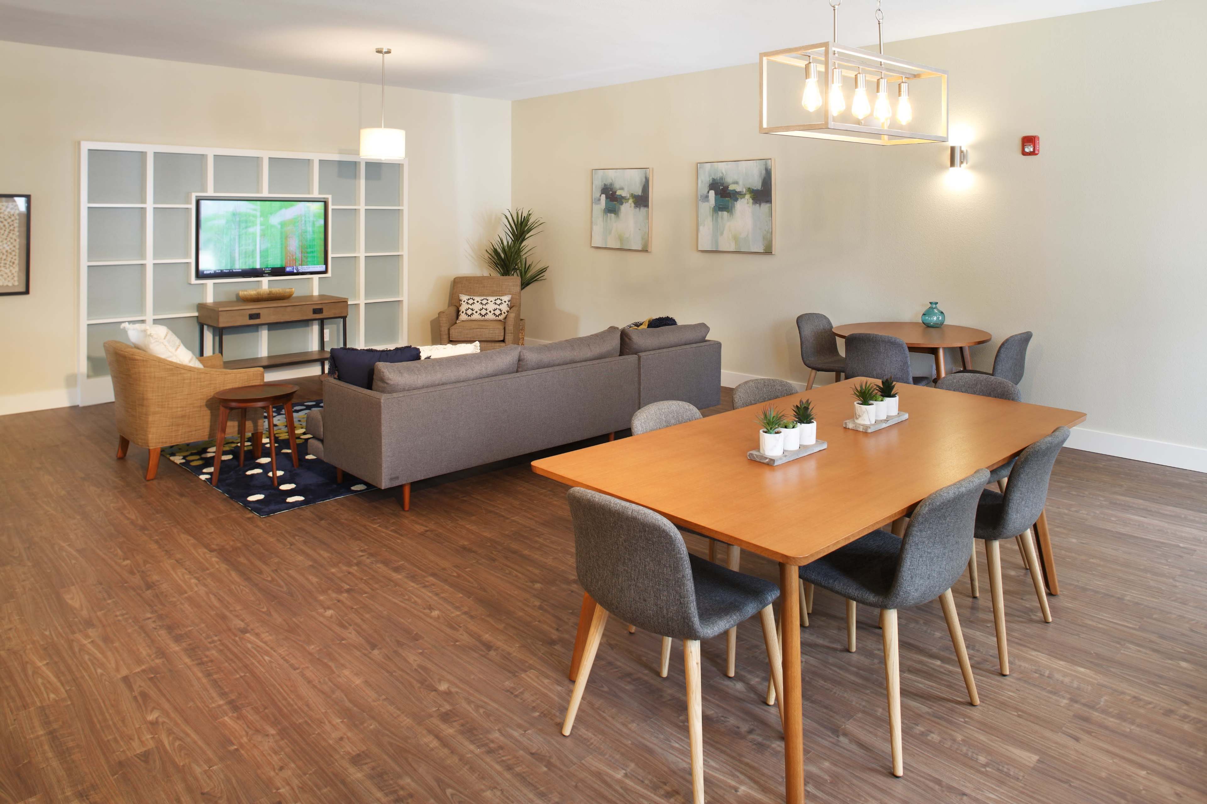 A modern living room features a sofa, coffee table, and television against one wall, with a dining table and chairs on the other side.