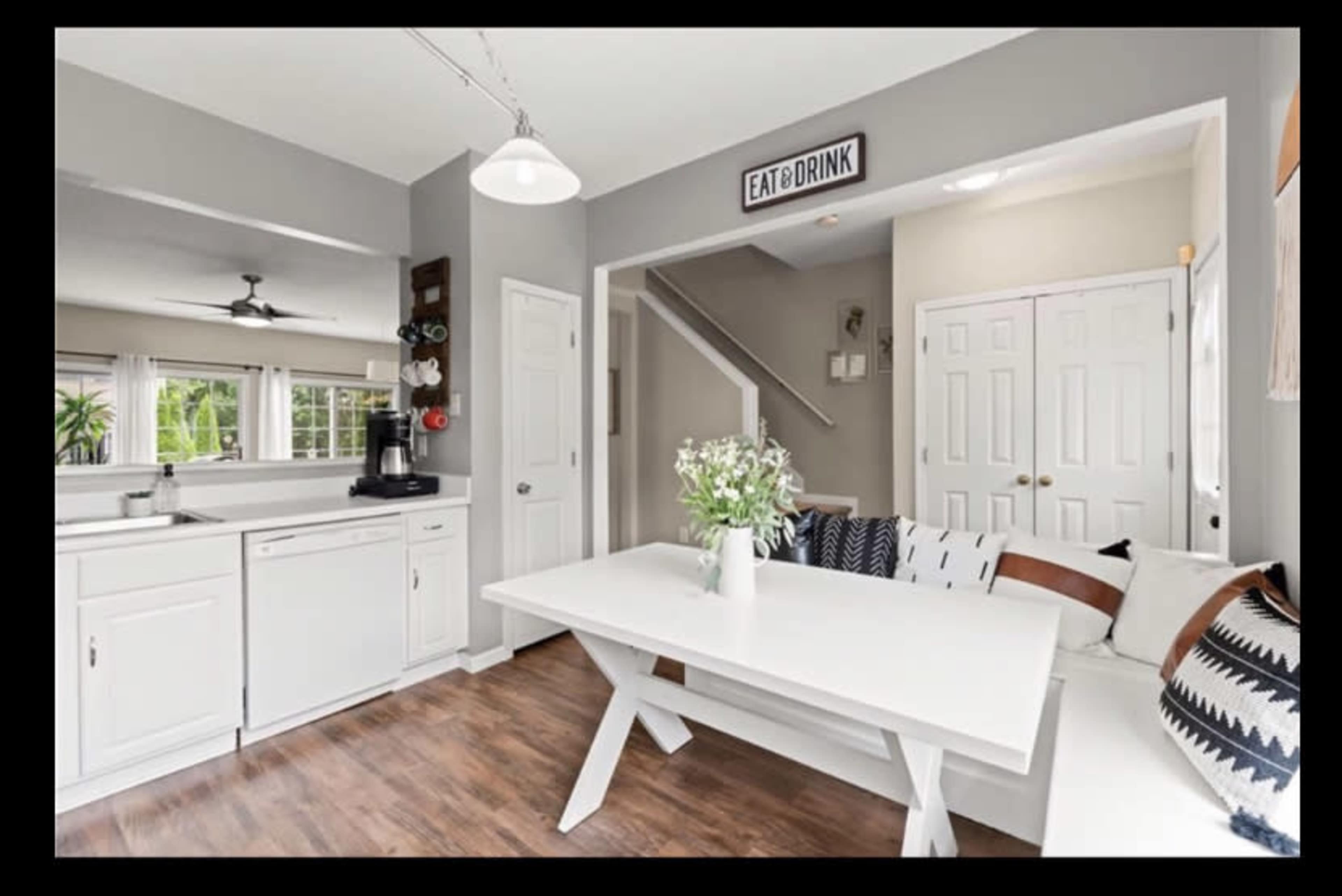 A modern kitchen features a white table, a seating area with decorative pillows, and a coffee station, all set in a light and airy space.