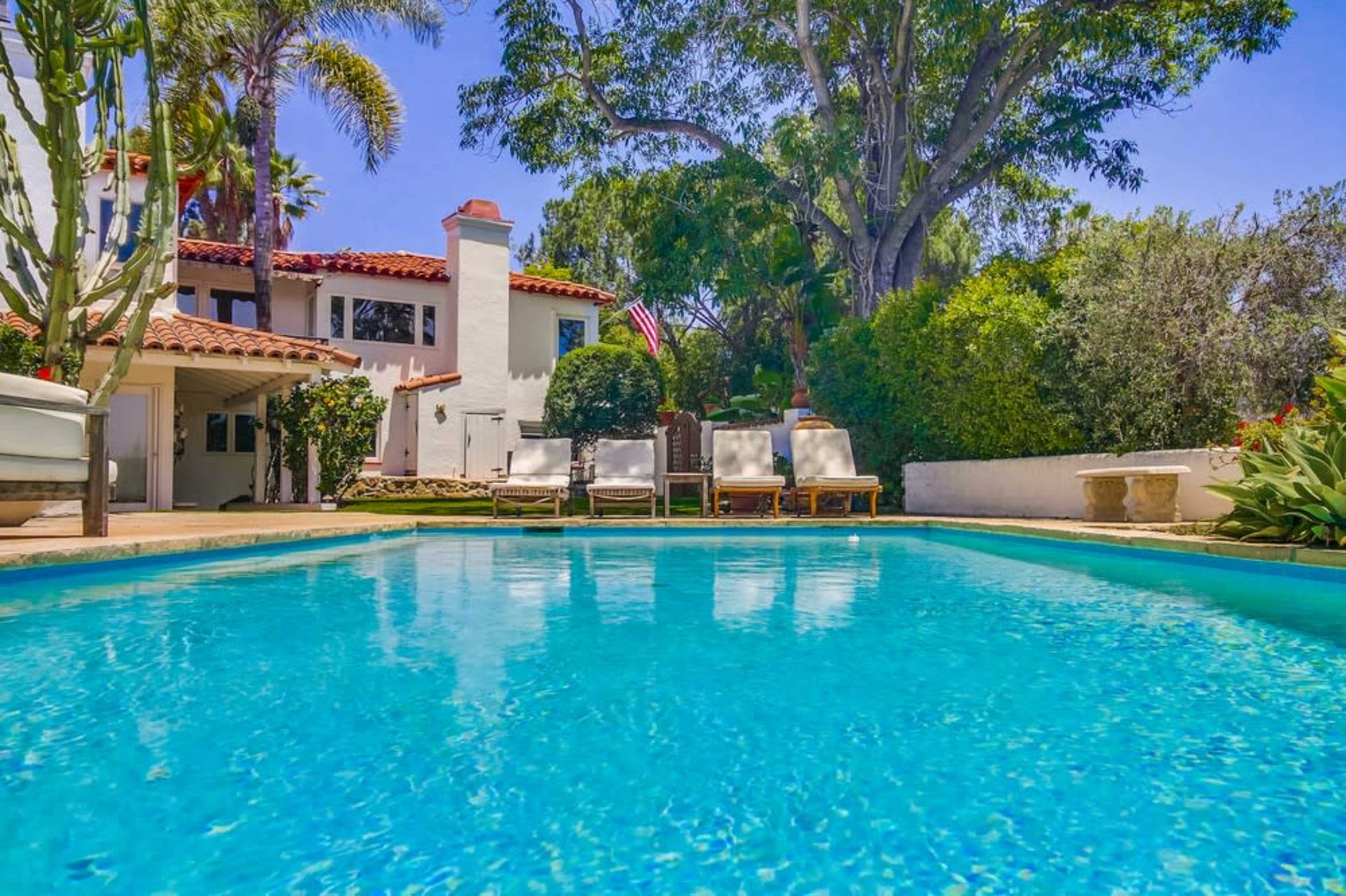 The scene features a blue swimming pool in the foreground, with a white house and several lounge chairs surrounded by lush greenery in the background.