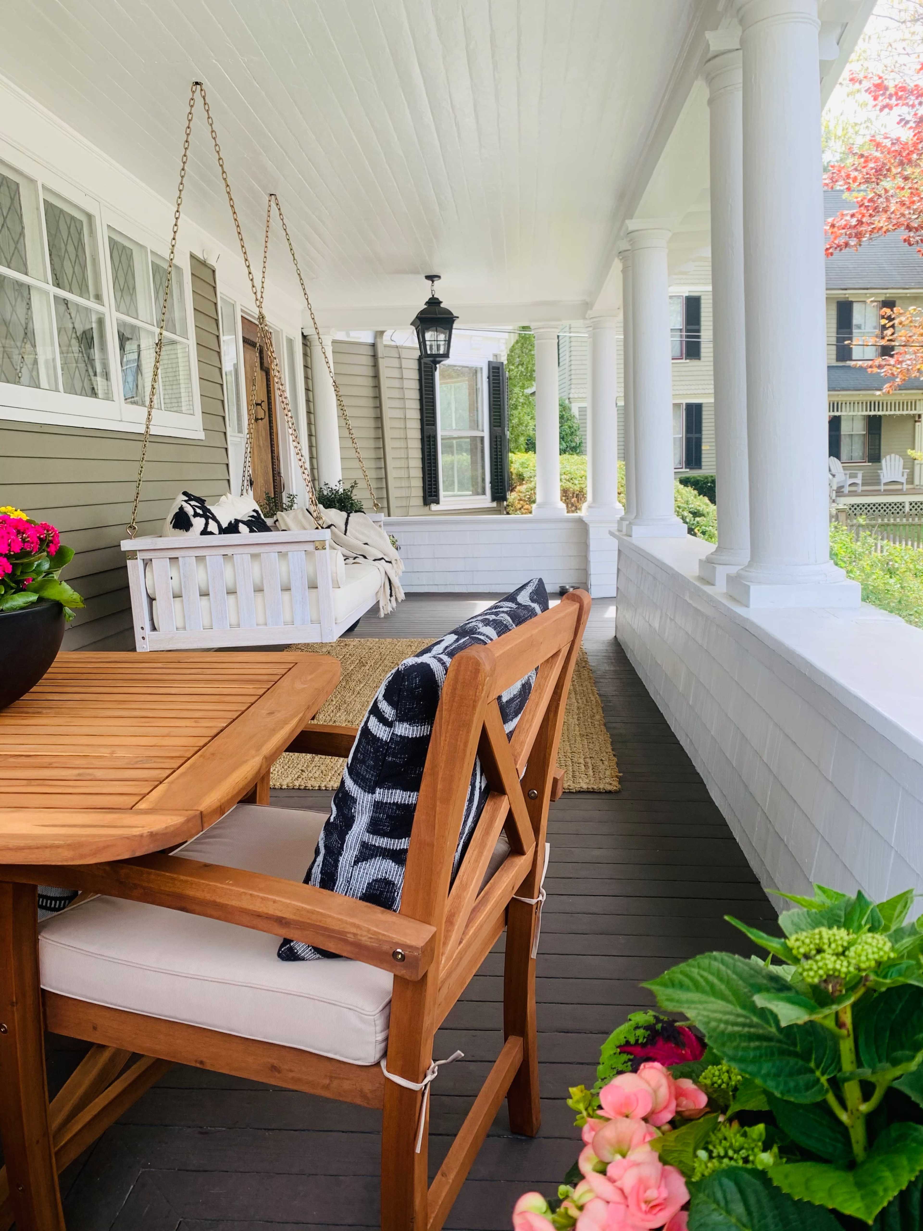 The image shows a front porch with a wooden dining table and chair, a swing hanging from the ceiling, and potted plants along the railing.