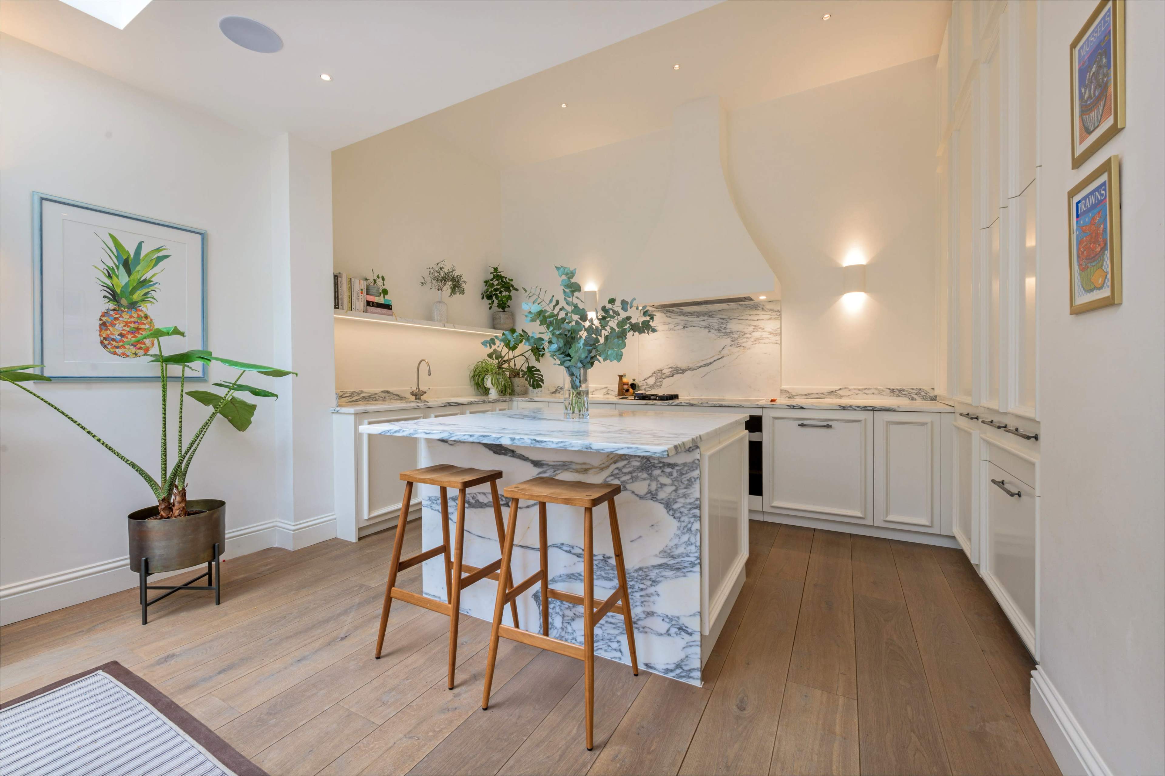 A modern kitchen features a marble island with two wooden stools, surrounded by white cabinets and minimal decorative elements.