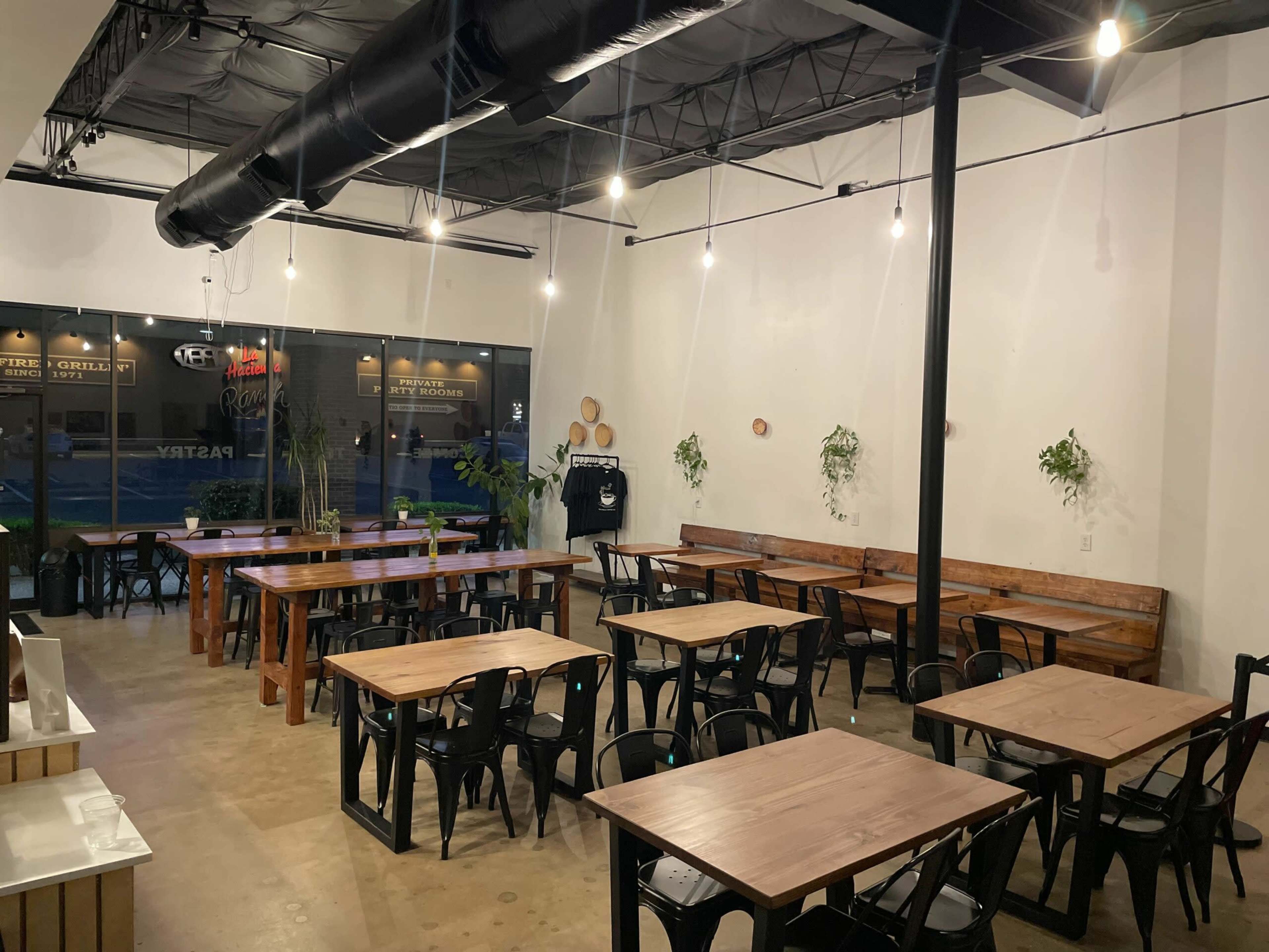The image shows a spacious, modern dining area with wooden tables, black metal chairs, and hanging light fixtures.