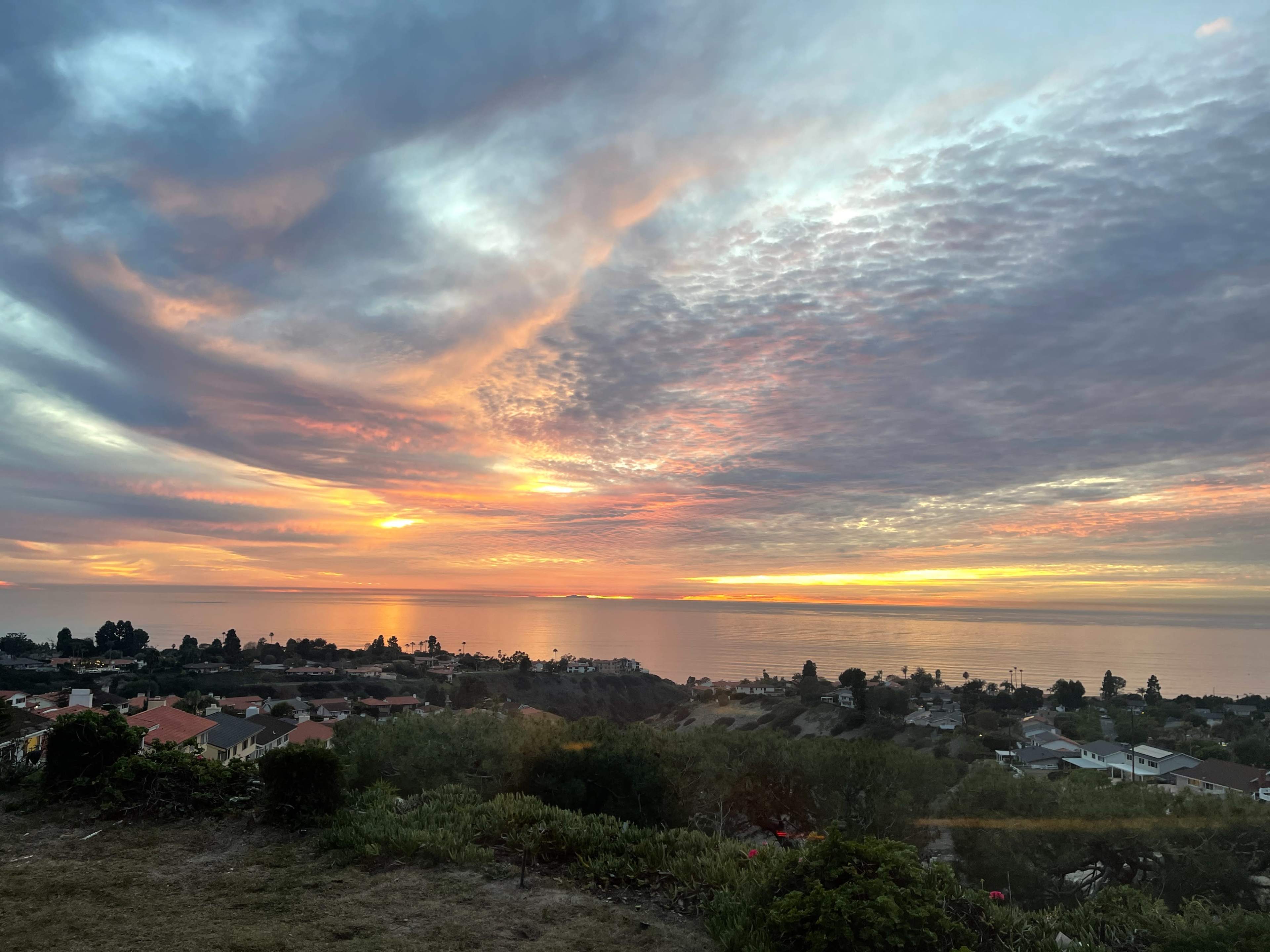 A vibrant sunset illuminates the sky over the ocean, casting colorful reflections on the water and highlighting the coastal landscape below.