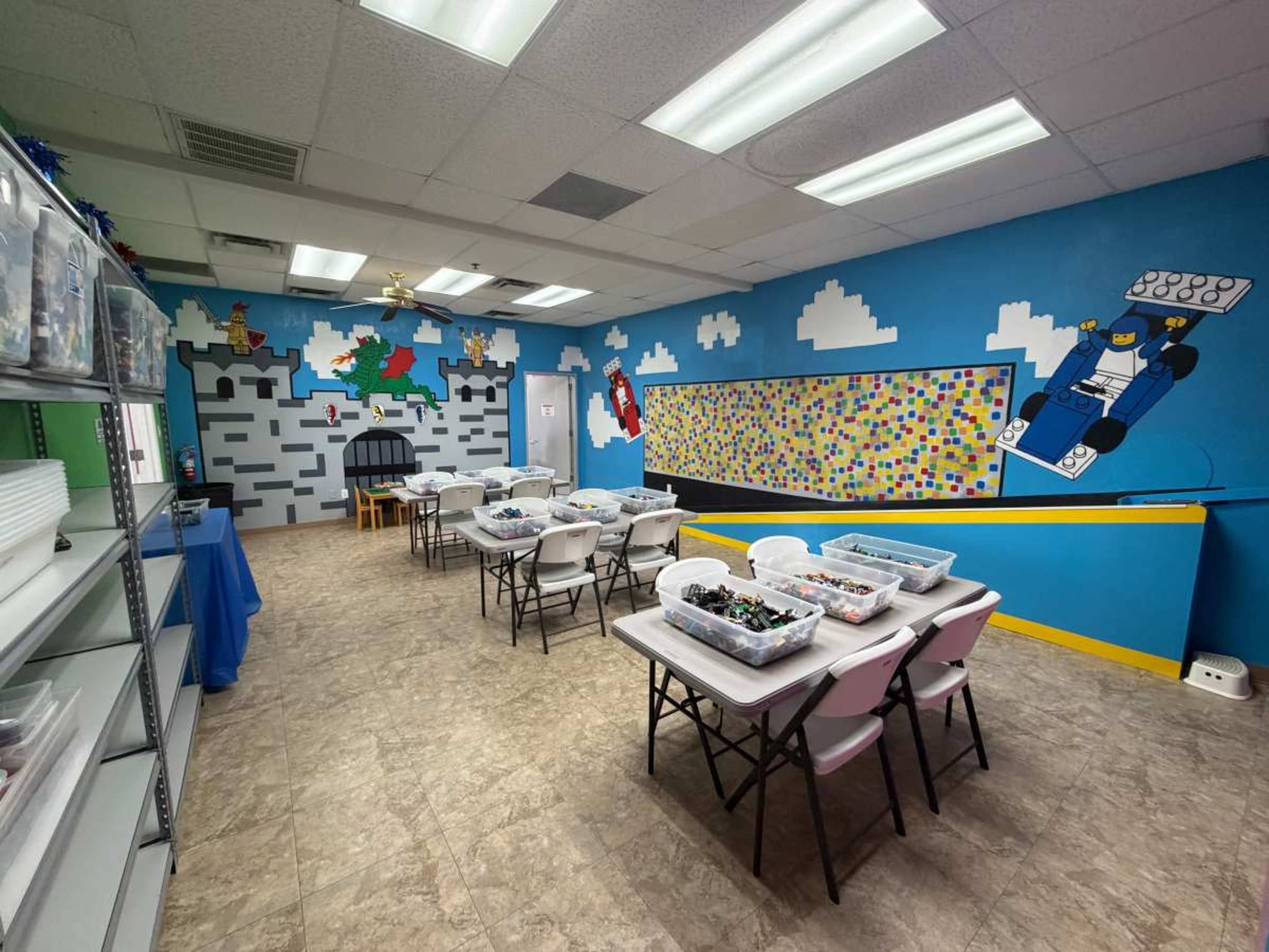 The image shows a bright classroom with several tables arranged for activities, surrounded by colorful murals of a castle and clouds on the walls.