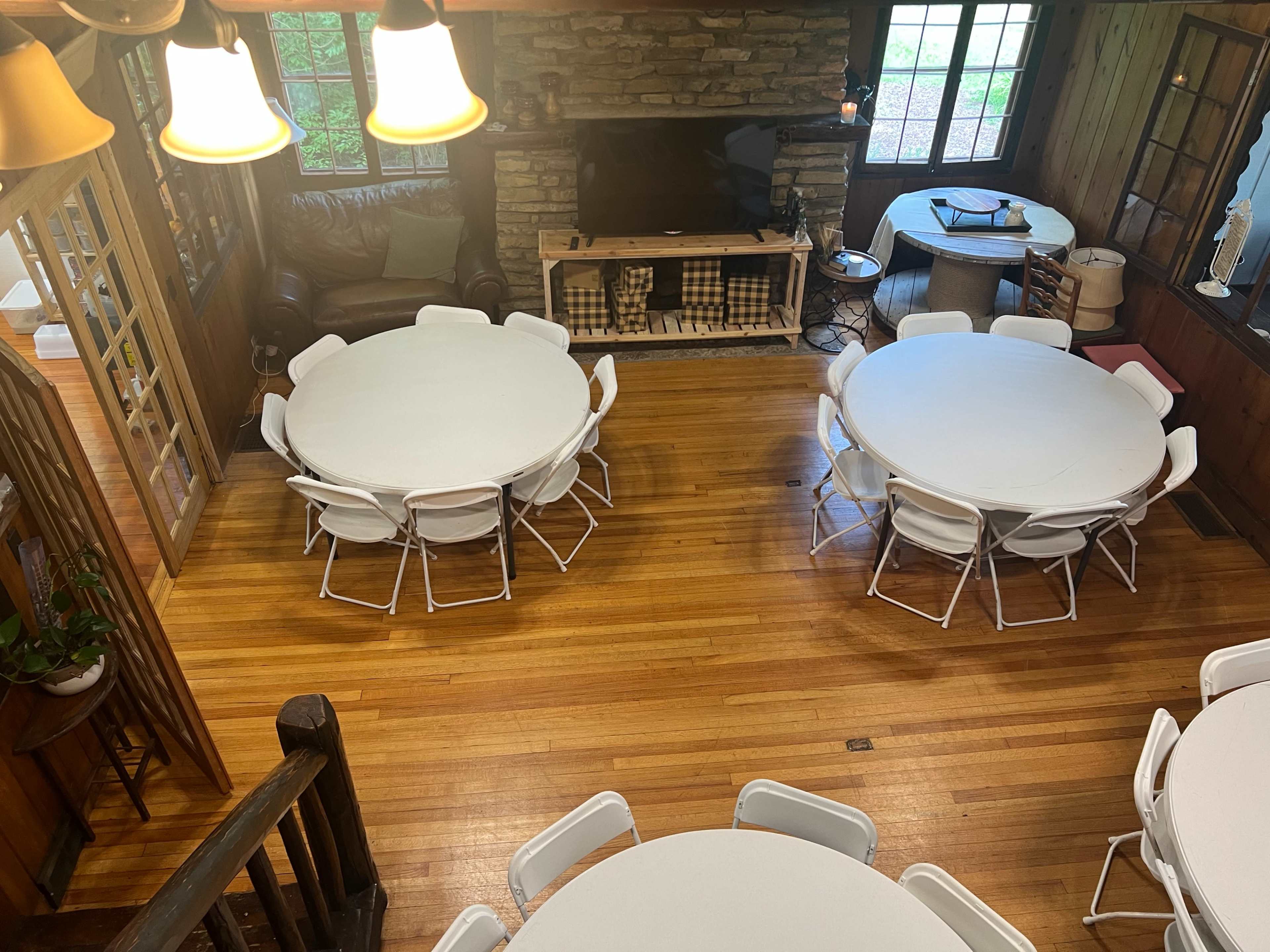 The image shows a spacious room with several round tables surrounded by white chairs, a fireplace against a stone wall, and large windows allowing natural light to enter.