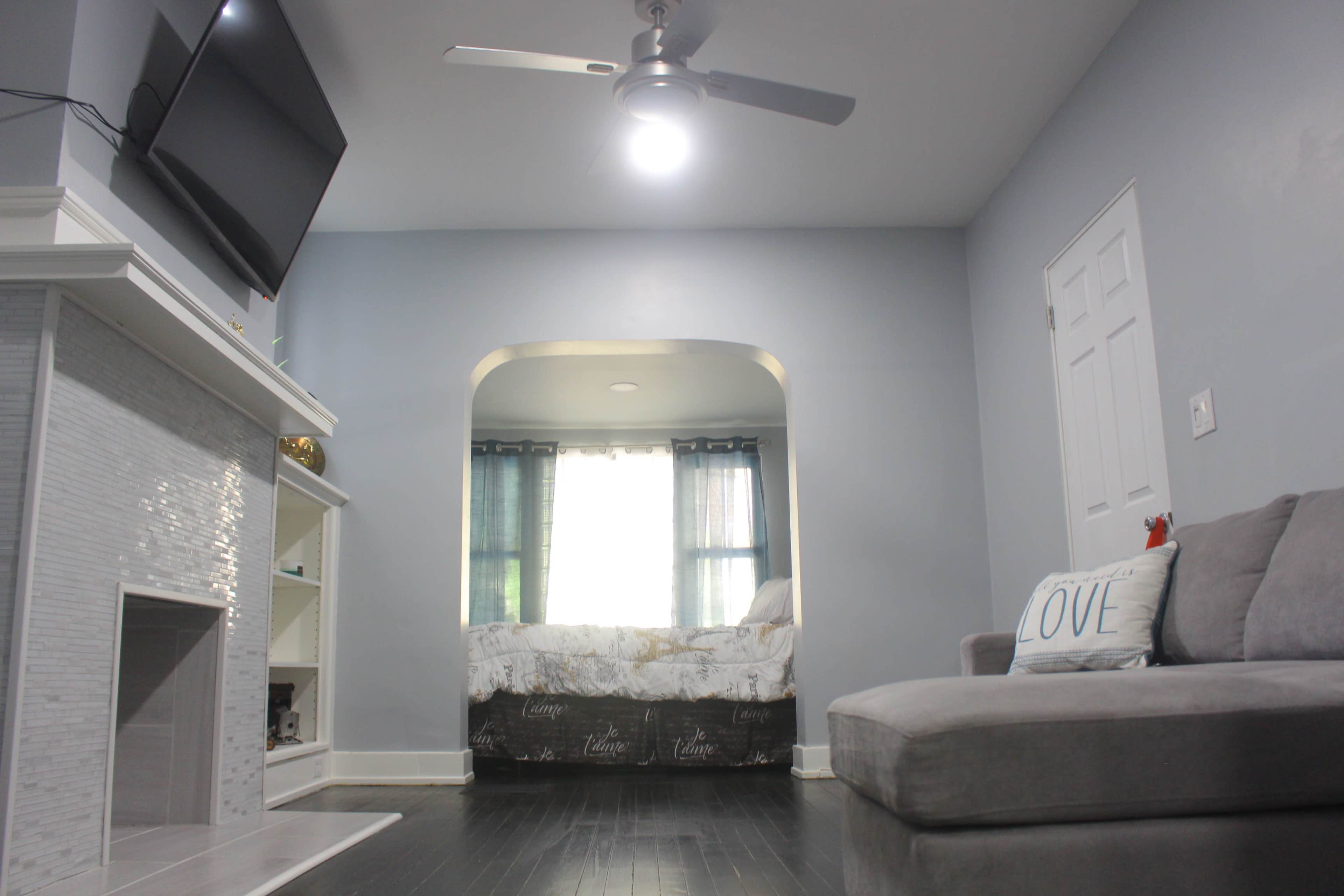 A living room with a light gray wall, a television mounted on the wall, a gray sectional sofa, and an archway leading to a bedroom with a bed and curtains.