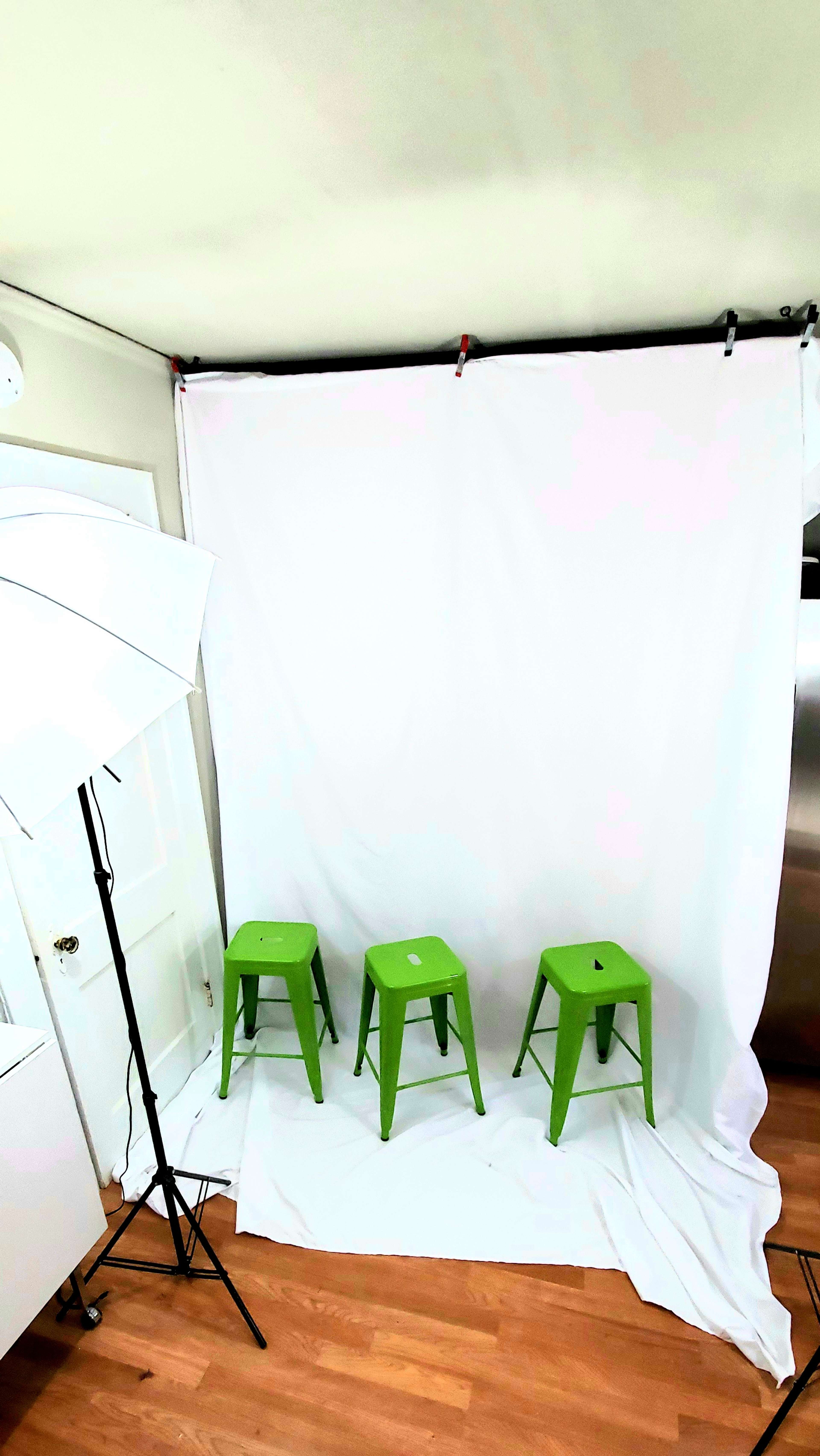 A photography setup with three green stools in front of a white backdrop, accompanied by a lighting umbrella and wooden flooring.