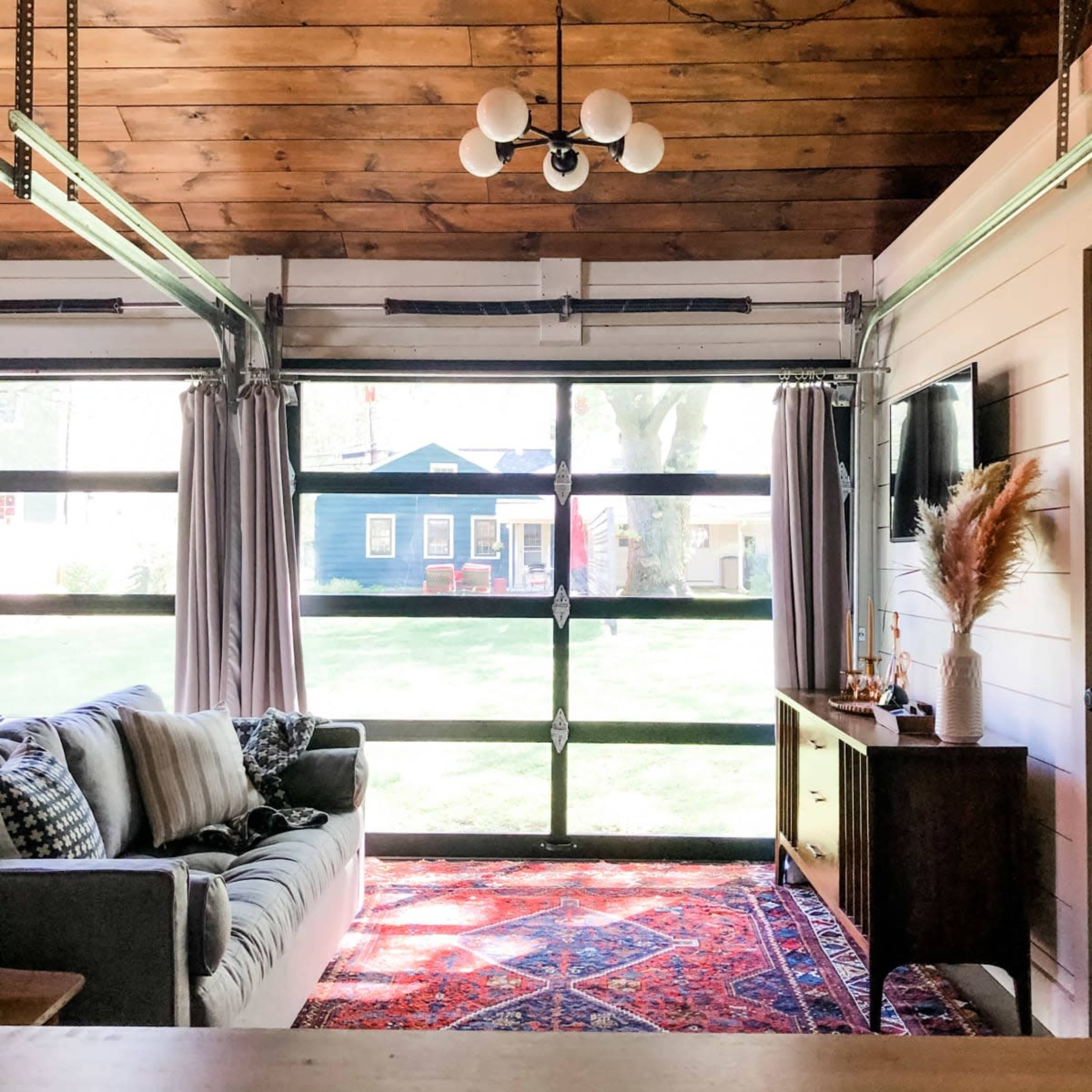 The image shows a cozy living space with a sofa, a TV, and a view of a yard through a large glass garage door.