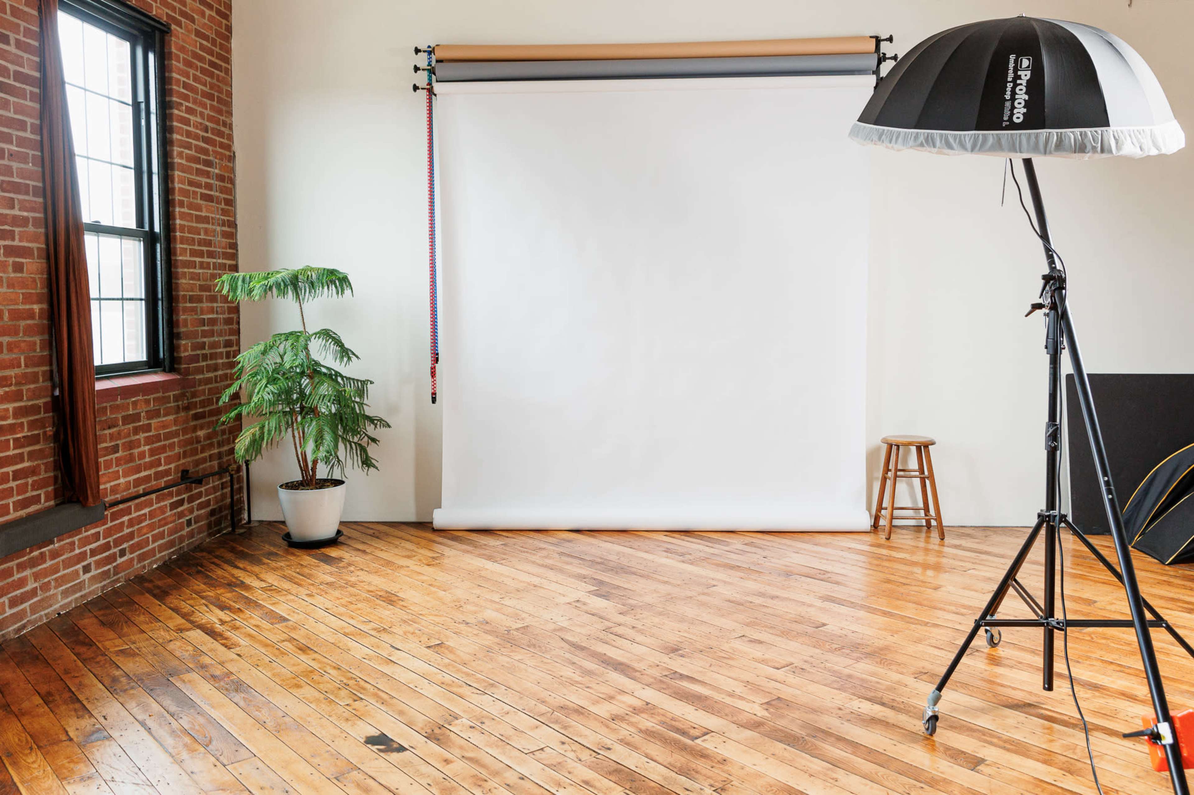 A photography studio featuring a smooth white backdrop, a tripod light with a softbox, a potted plant, a wooden stool, and exposed brick walls.