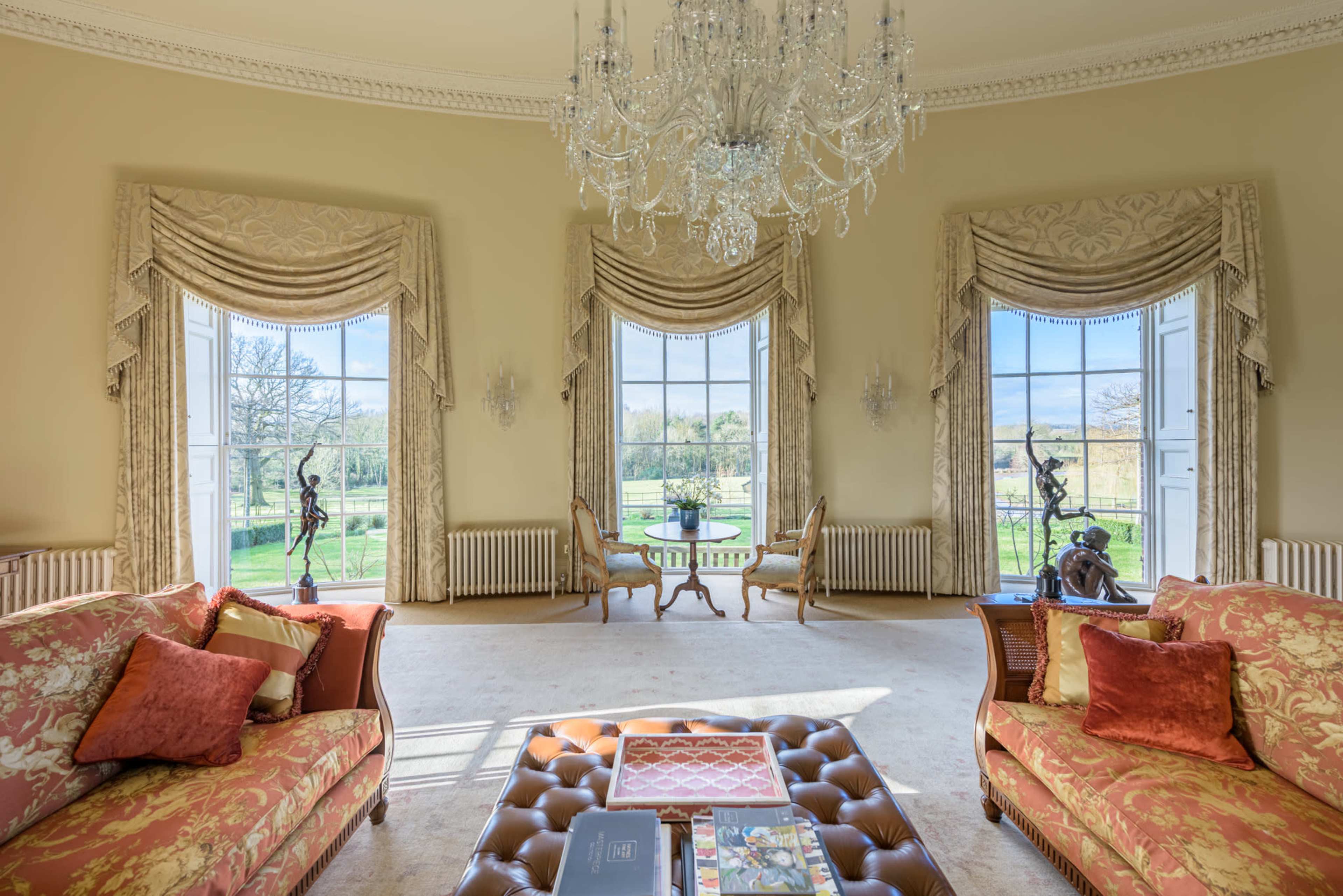 The image shows an elegantly furnished room with large windows showcasing a view of a garden and ornate decor including a chandelier and floral-patterned sofas.