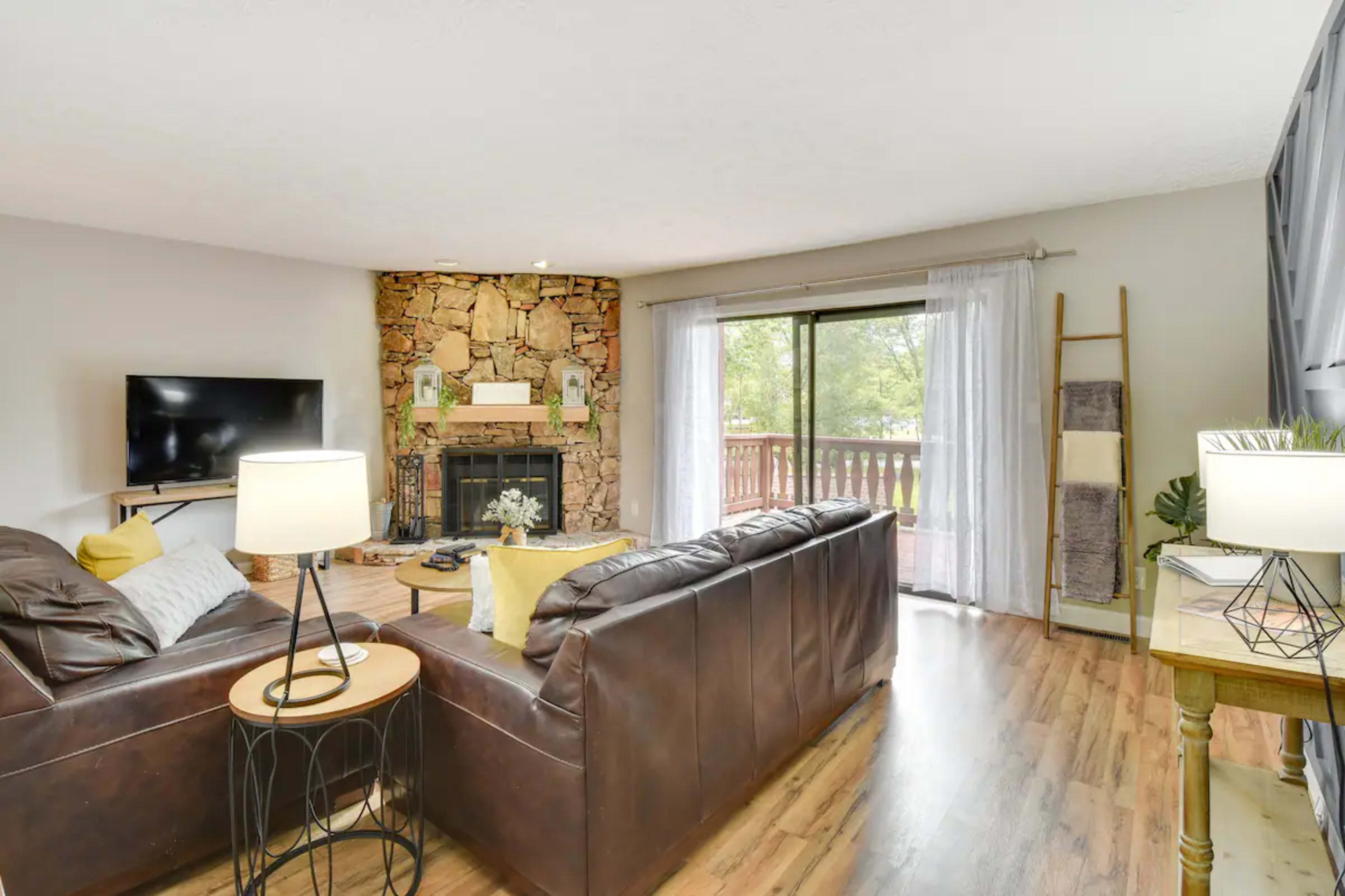 A living room with a stone fireplace, a dark leather sofa, a television, and large windows that open to a view of the outdoors.