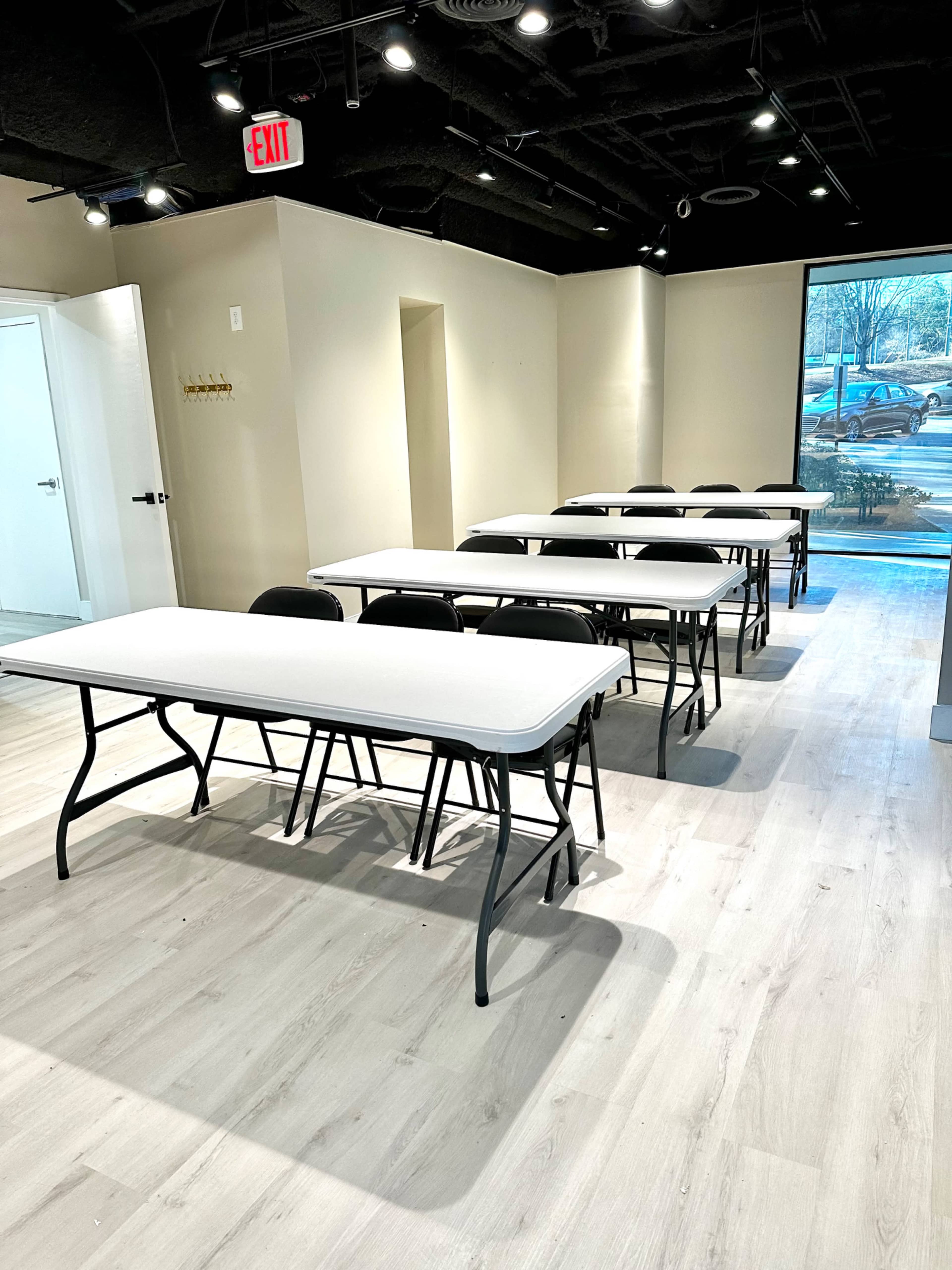 A classroom or meeting space arranged with several white folding tables and black chairs facing forward, illuminated by overhead lights.