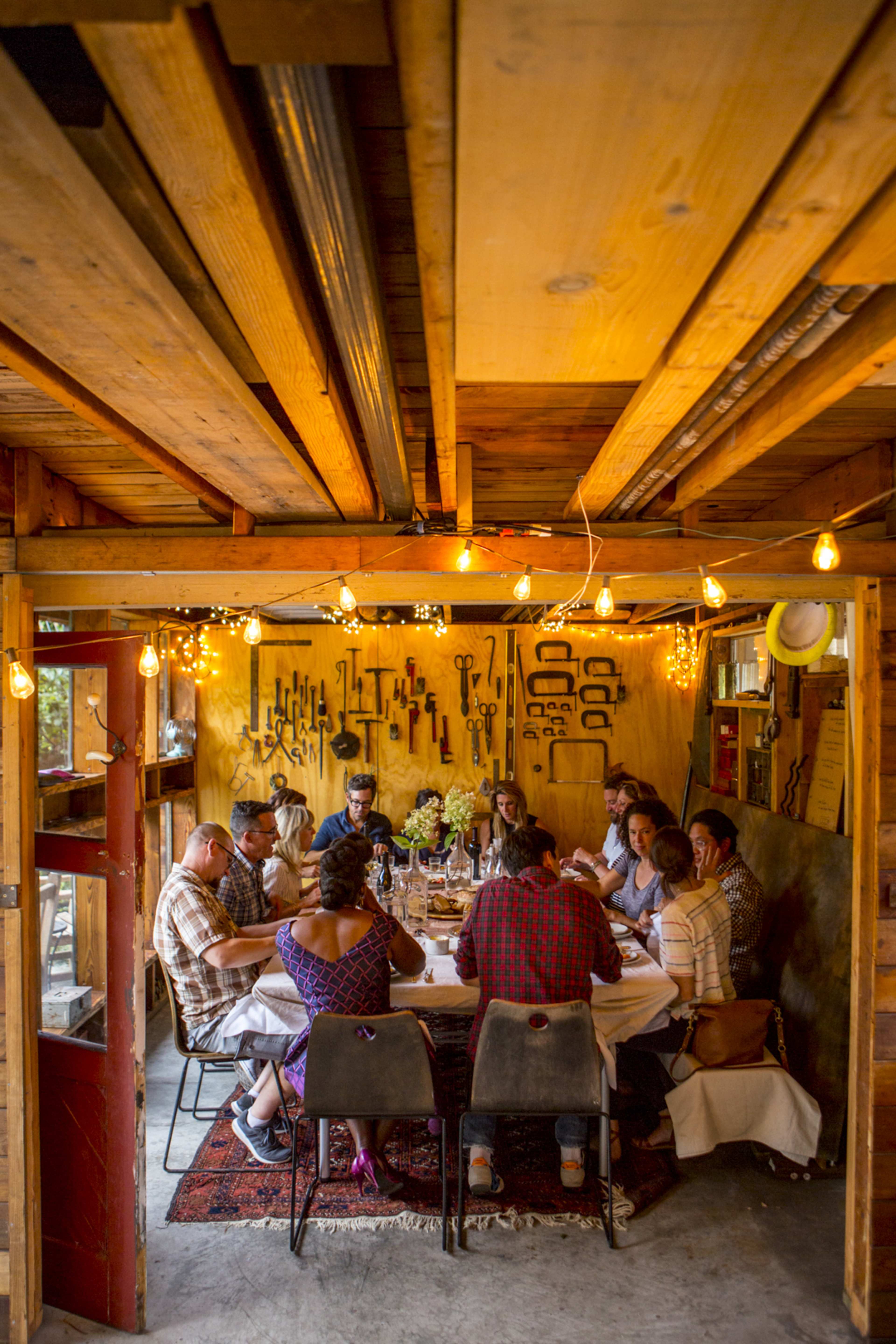 A group of people sits around a long table in a rustic dining space, illuminated by string lights, with tools displayed on the wall behind them.