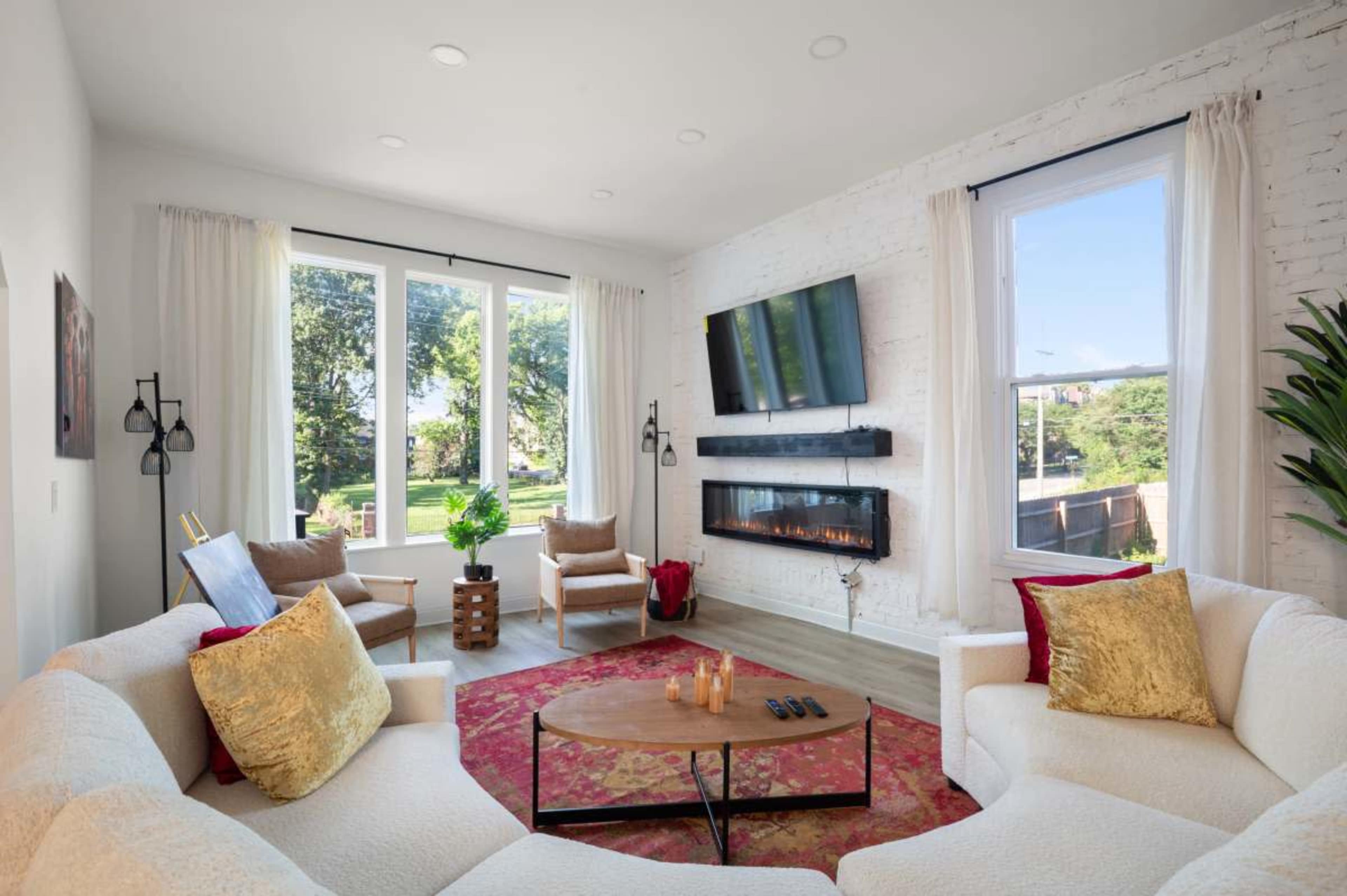 A modern living room features a white sofa, a coffee table, and a wall-mounted TV above a sleek electric fireplace, with large windows allowing natural light and greenery outside.
