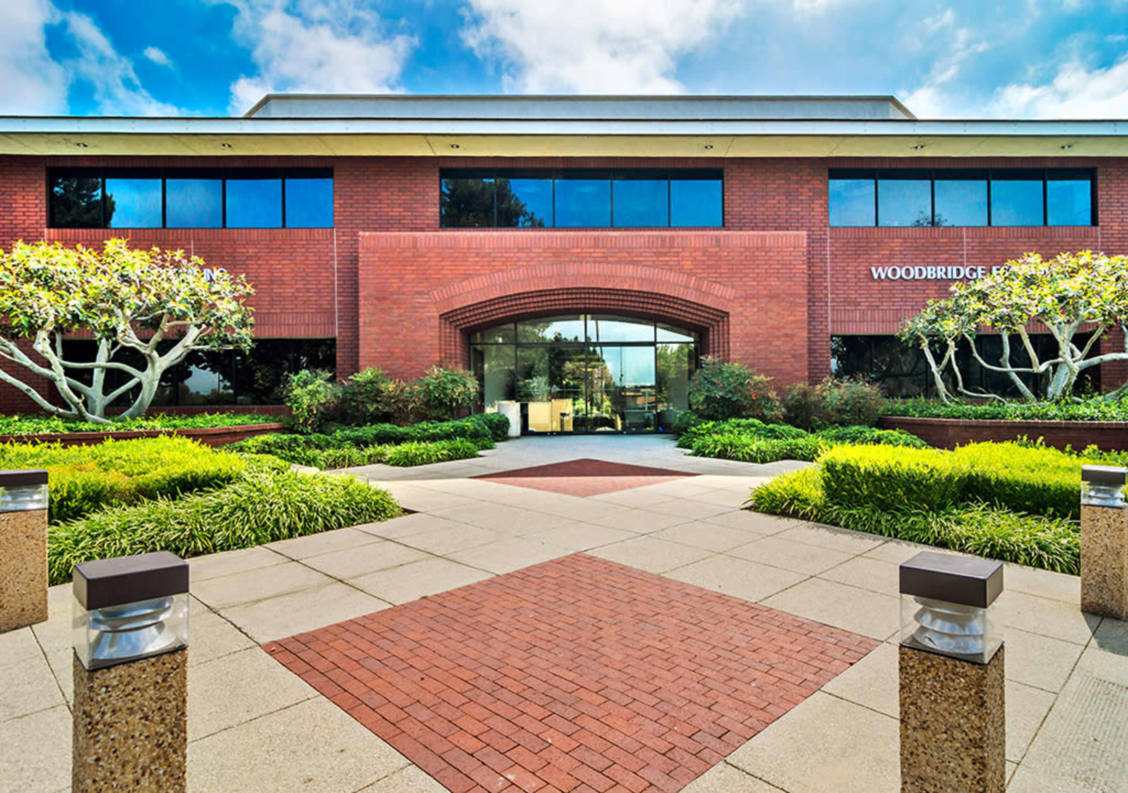 The image shows a red brick office building with large windows, surrounded by landscaped greenery and a paved pathway leading to the entrance.