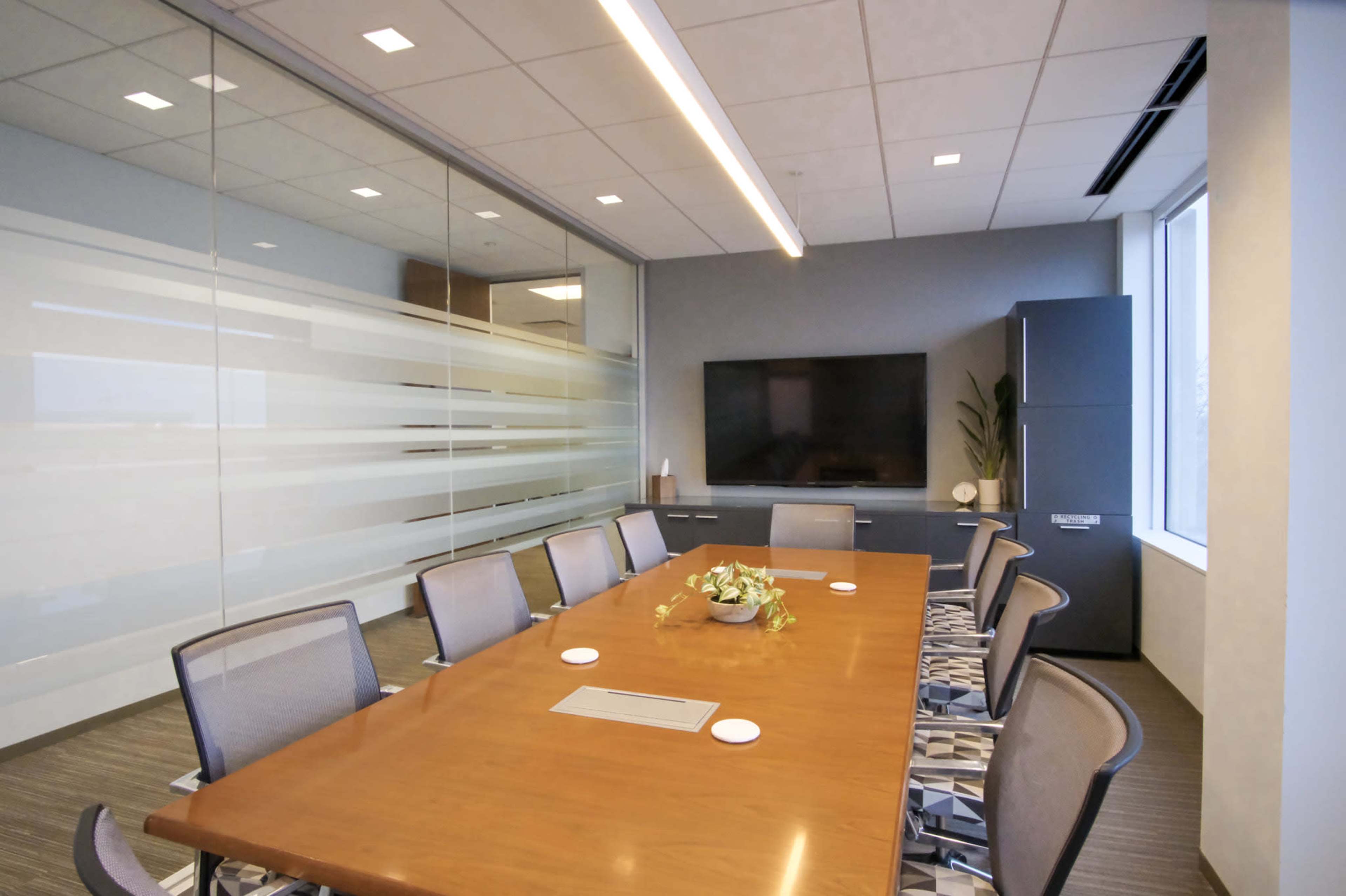 The image shows a modern conference room with a large wooden table, several ergonomic chairs, a wall-mounted television, and a floor-to-ceiling glass partition.