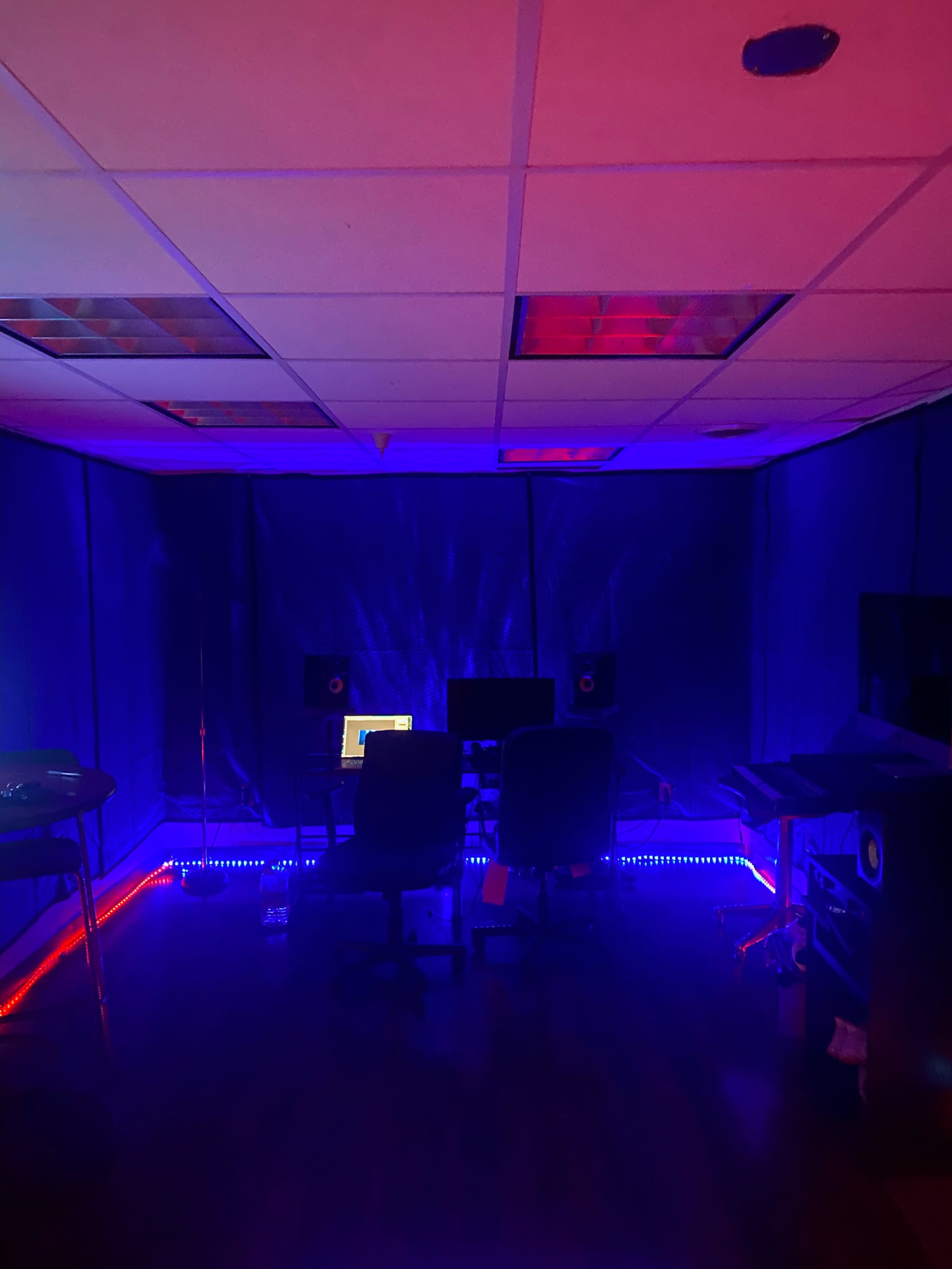 A dimly lit studio room with blue and red lighting, featuring two chairs and multiple computer screens set up on desks.