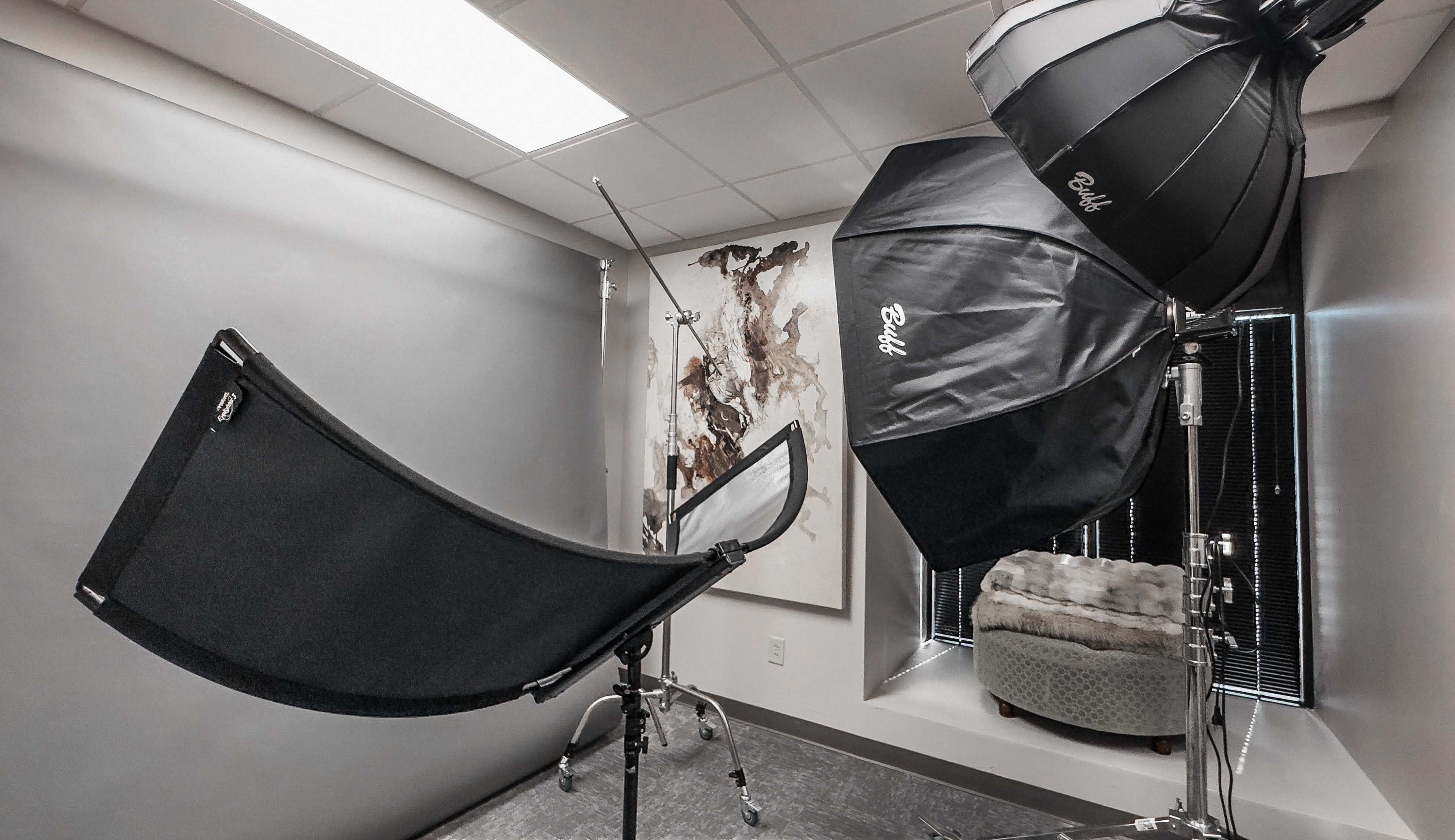 The image shows a photography studio setup featuring softbox lighting, a backdrop stand with a draped cloth, and a comfortable chair in the corner.