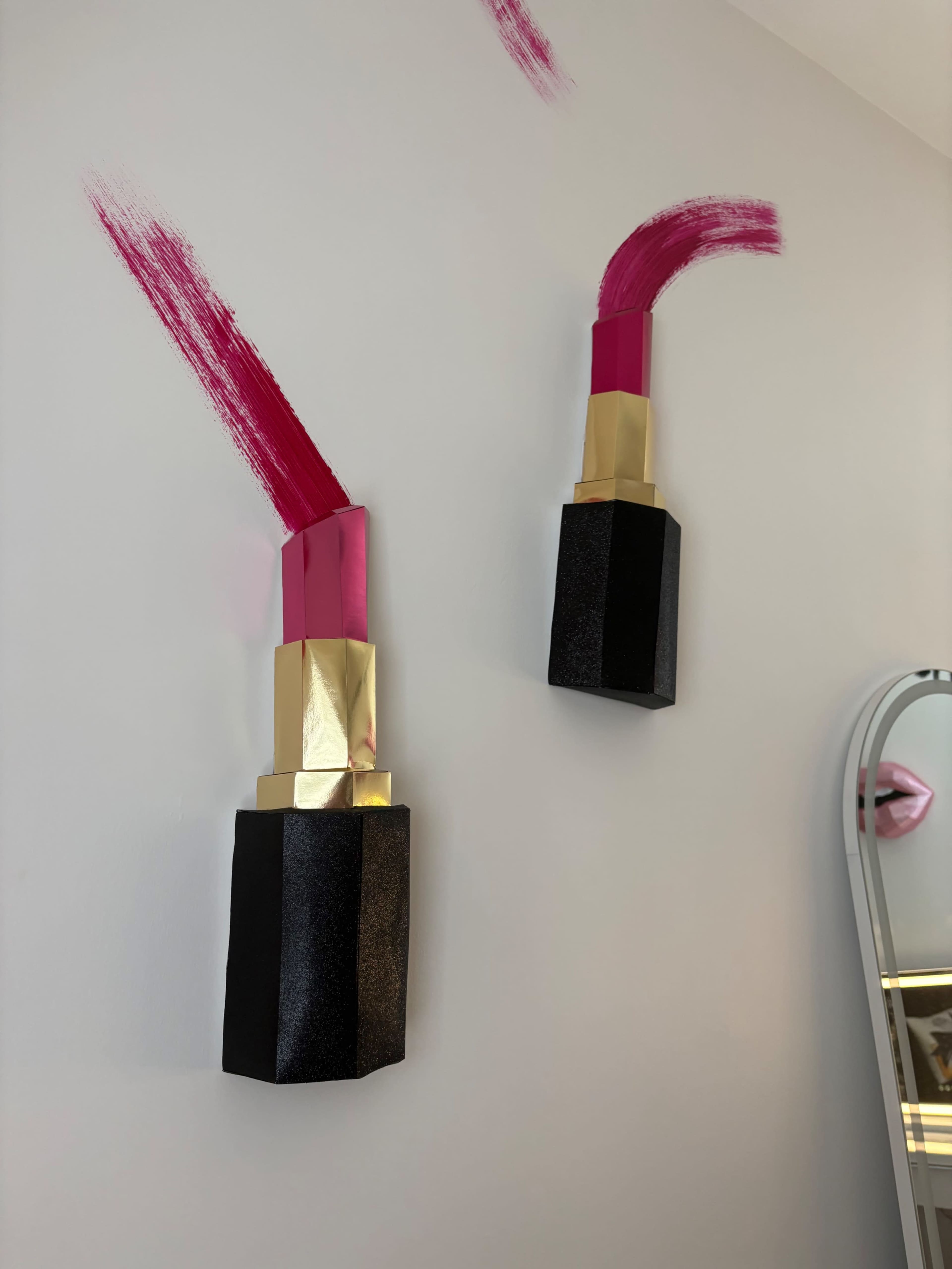 Two large, decorative lipstick sculptures are mounted on a wall, alongside bold pink brush strokes.