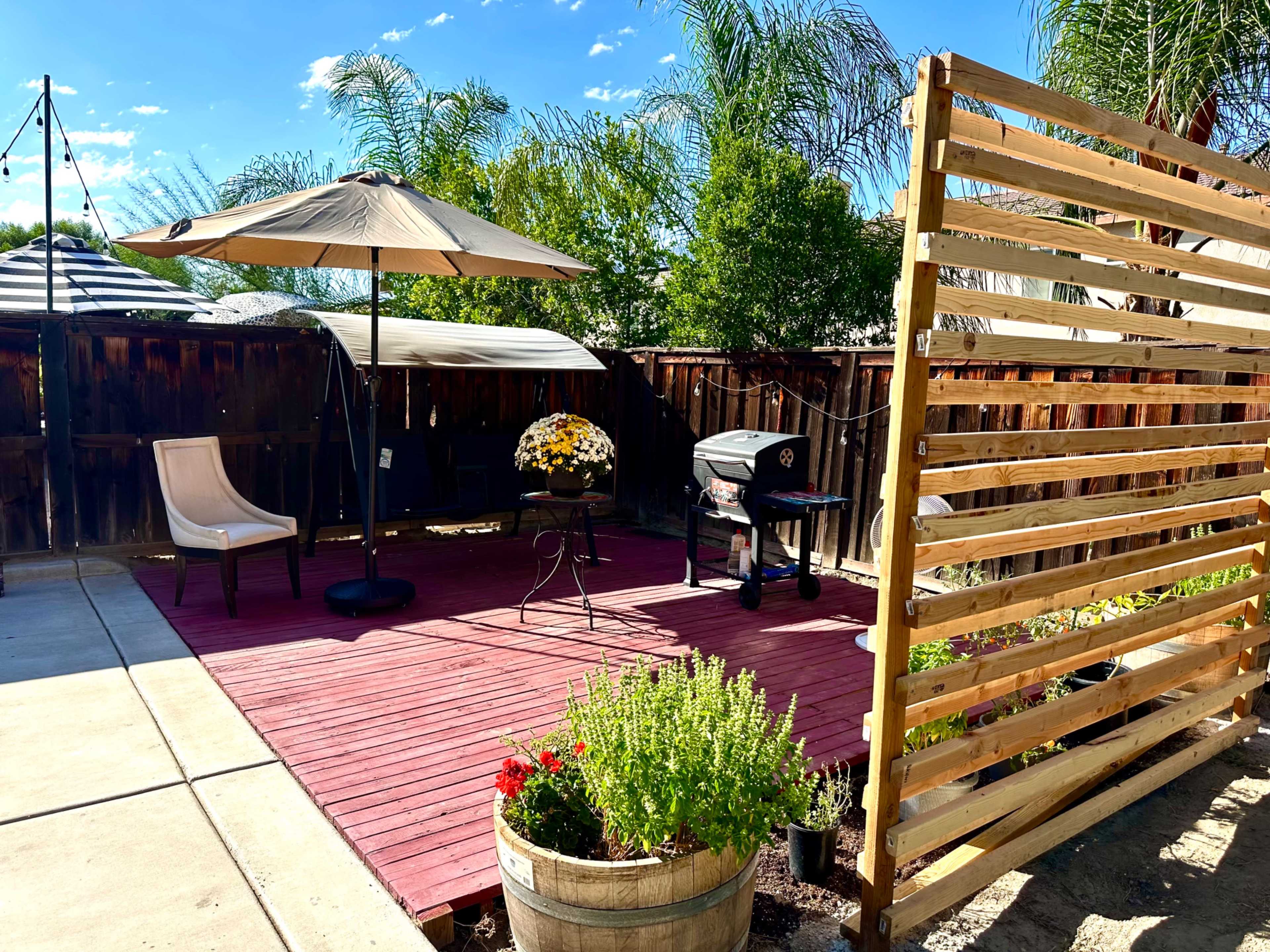 A wooden deck area features a patio umbrella, seating, a small table, a grill, and potted plants surrounded by a wooden privacy screen.