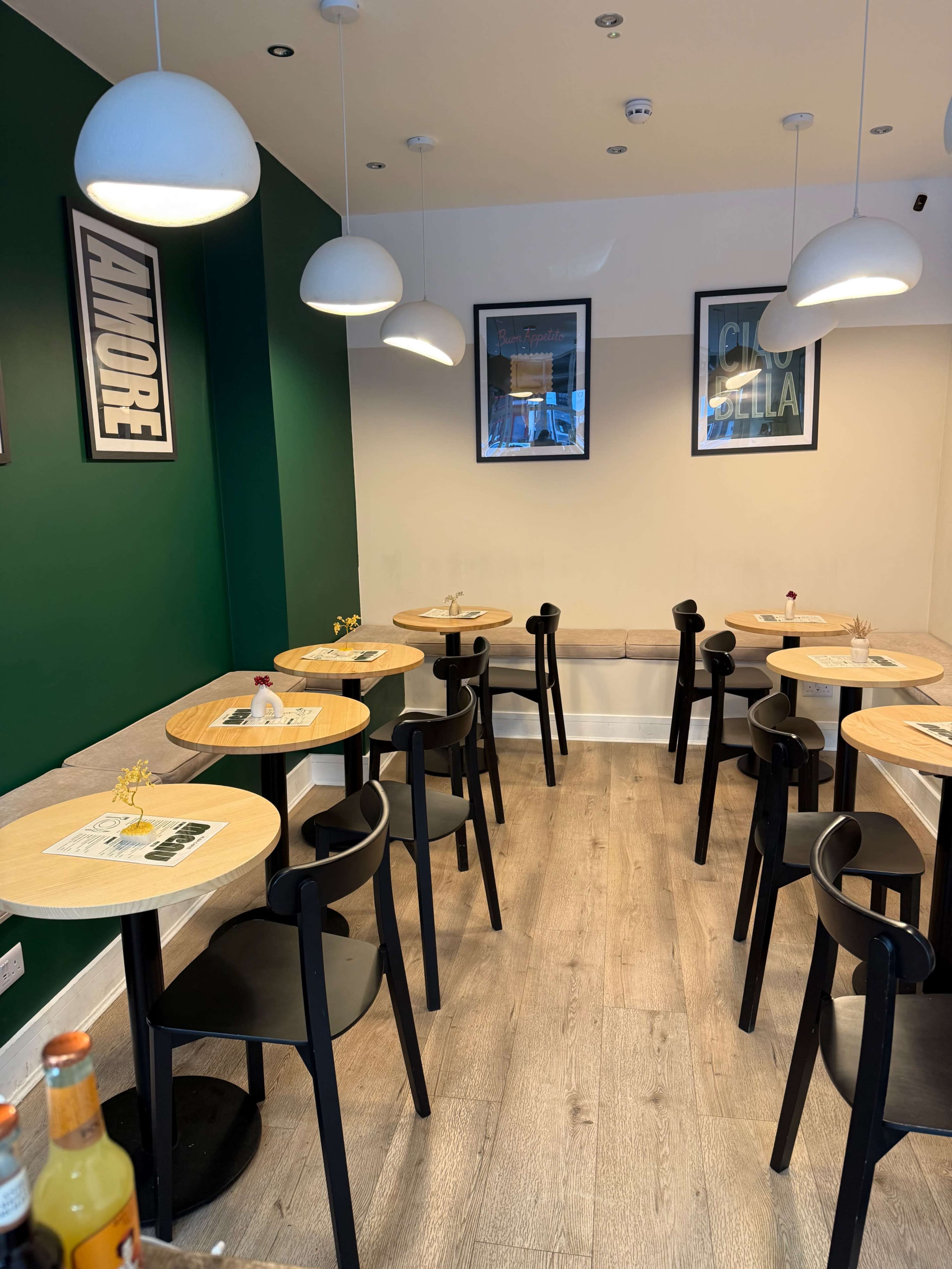 The image shows a modern restaurant interior with multiple round wooden tables and black chairs, surrounded by green and white walls and pendant lighting.