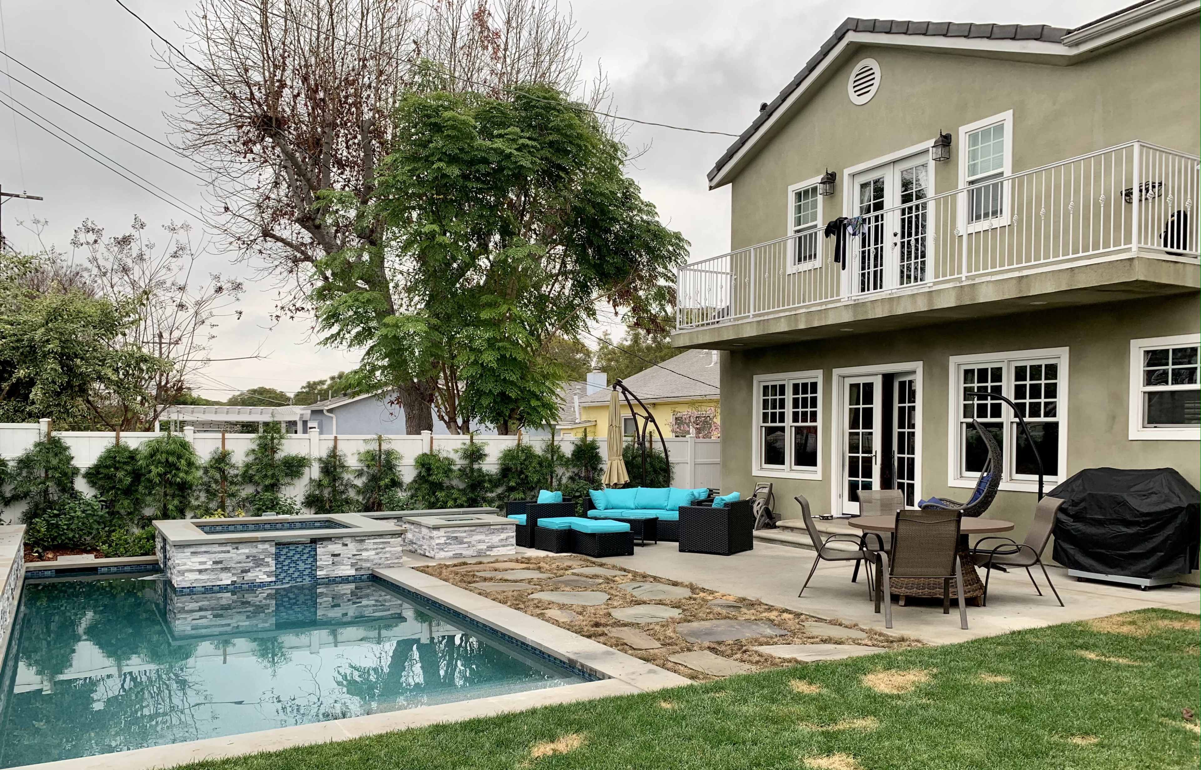 A modern backyard features a swimming pool, a seating area with turquoise furniture, and a two-story house with a balcony.