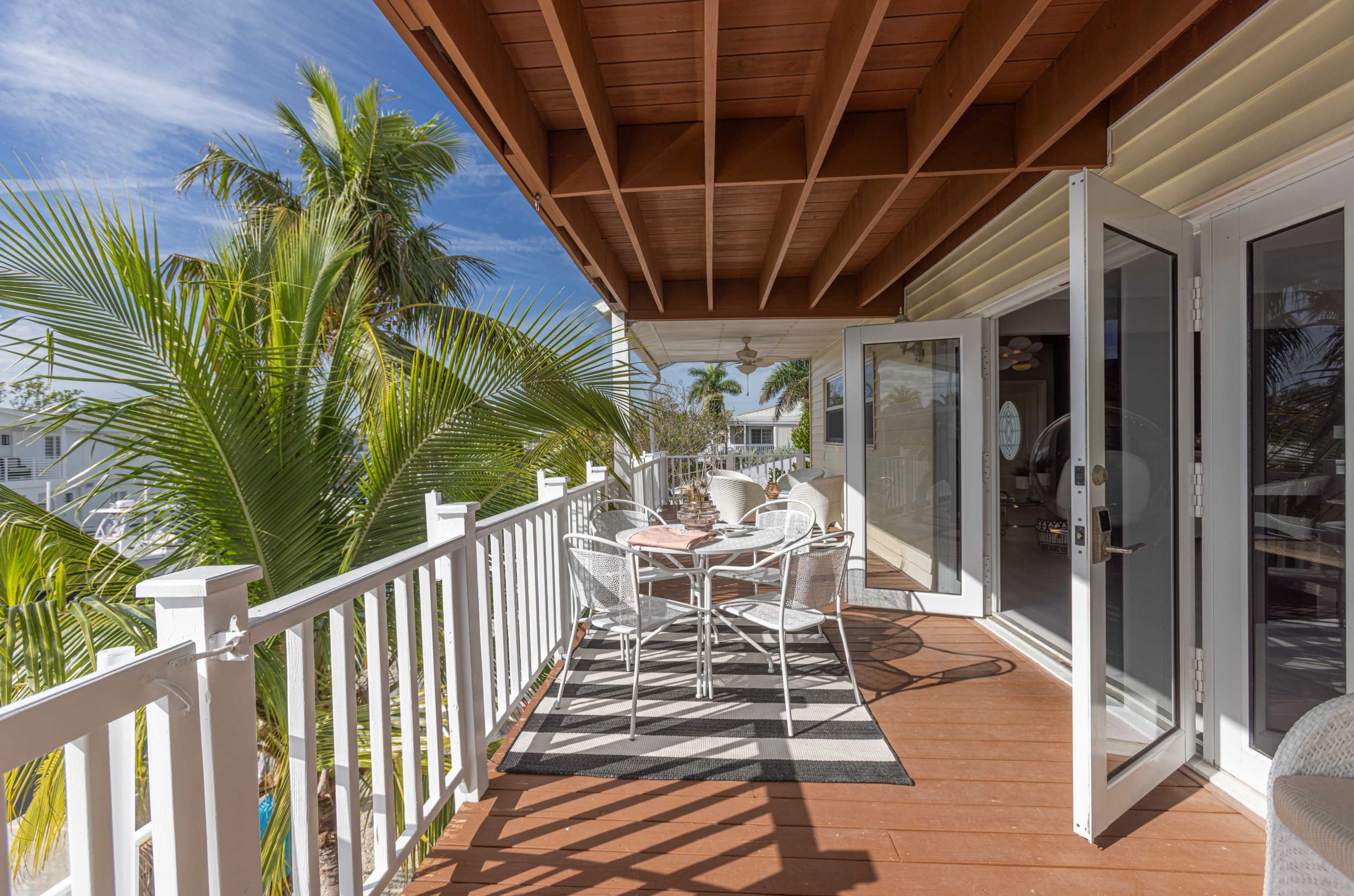 A wooden balcony features a dining set surrounded by tropical plants, with glass doors opened to an interior space.