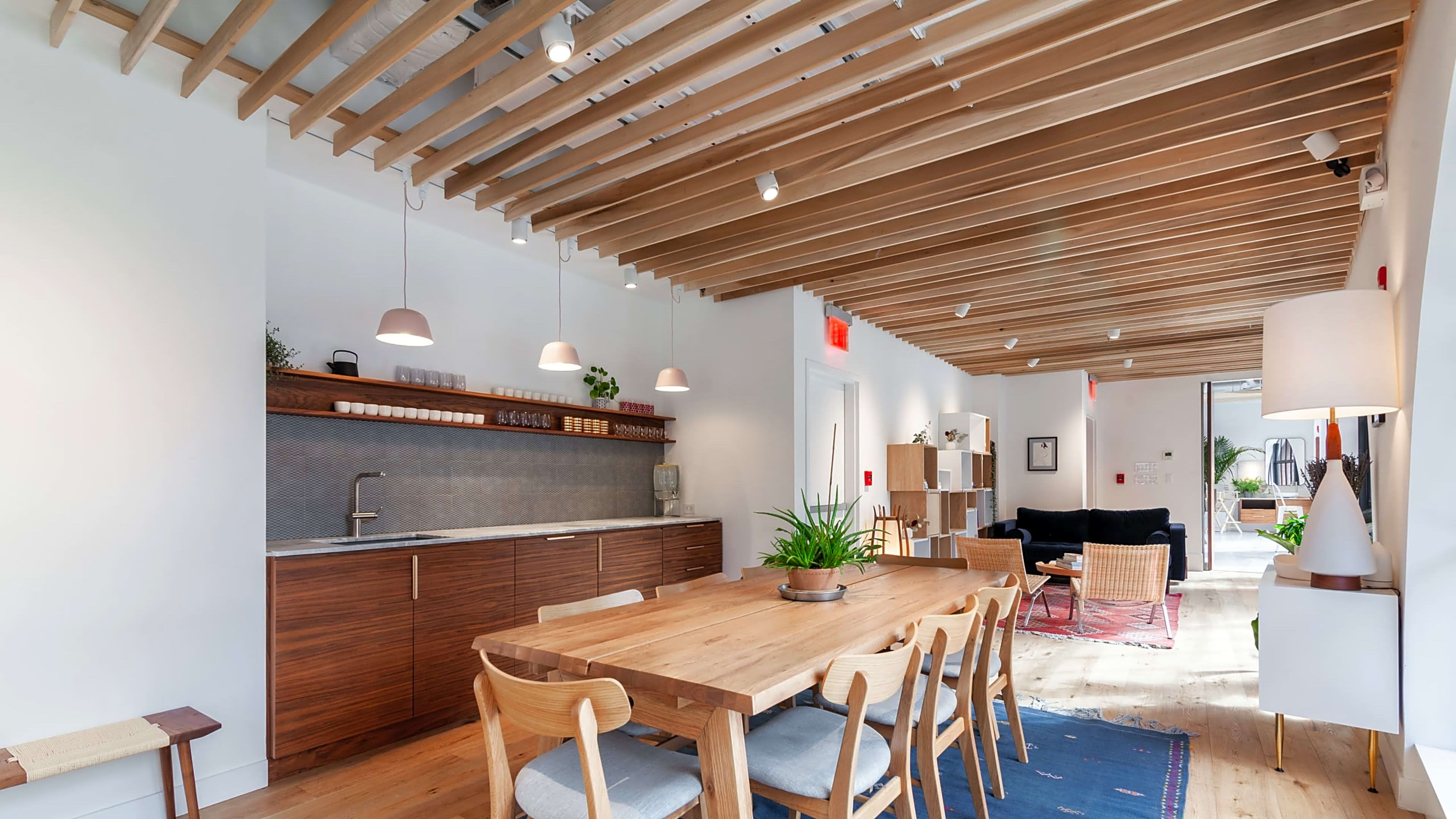 A modern dining area with a wooden table, minimalist chairs, and a kitchen along one wall featuring overhead lighting and a textured backsplash.